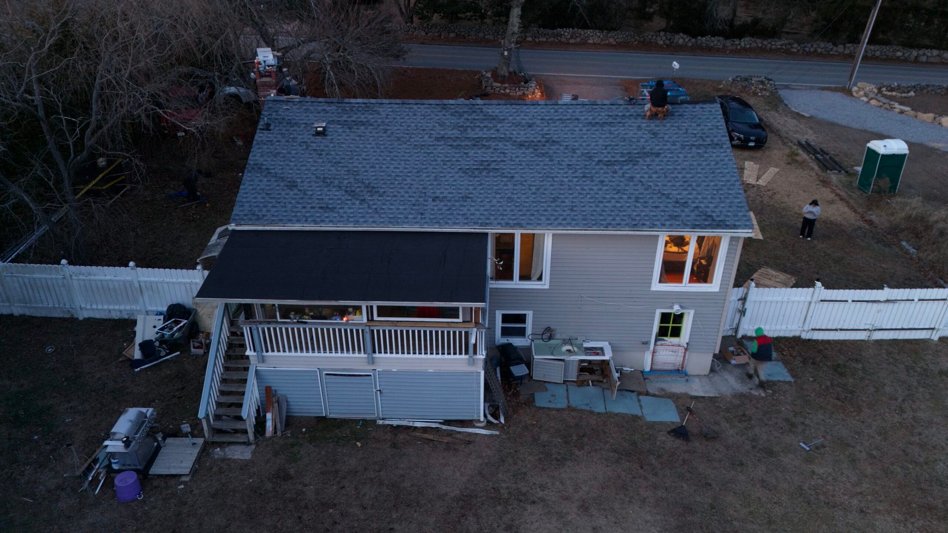 Aerial view of a small house with a covered porch and deck, surrounded by a white fence and bare trees.