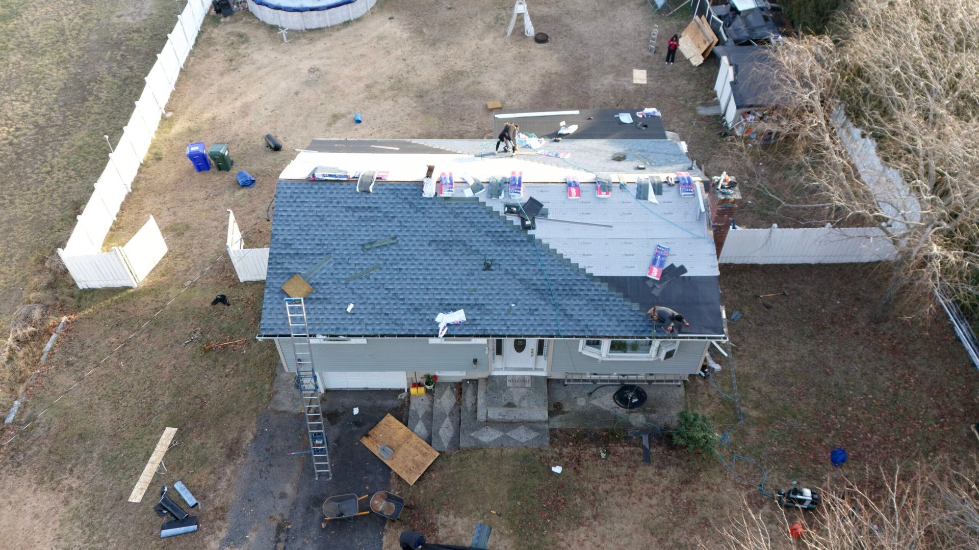 Aerial view of a house with a blue tarp roof and construction materials scattered around the yard.
