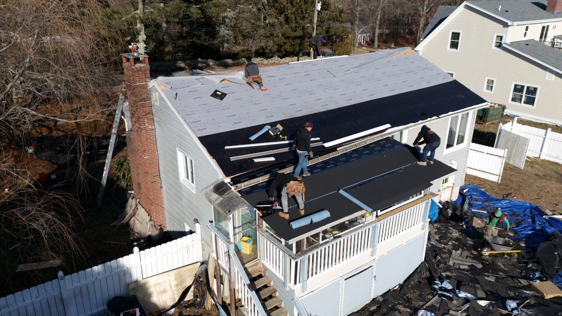 Workers installing a black roof on a house with a white deck and chimney, surrounded by trees and neighboring homes