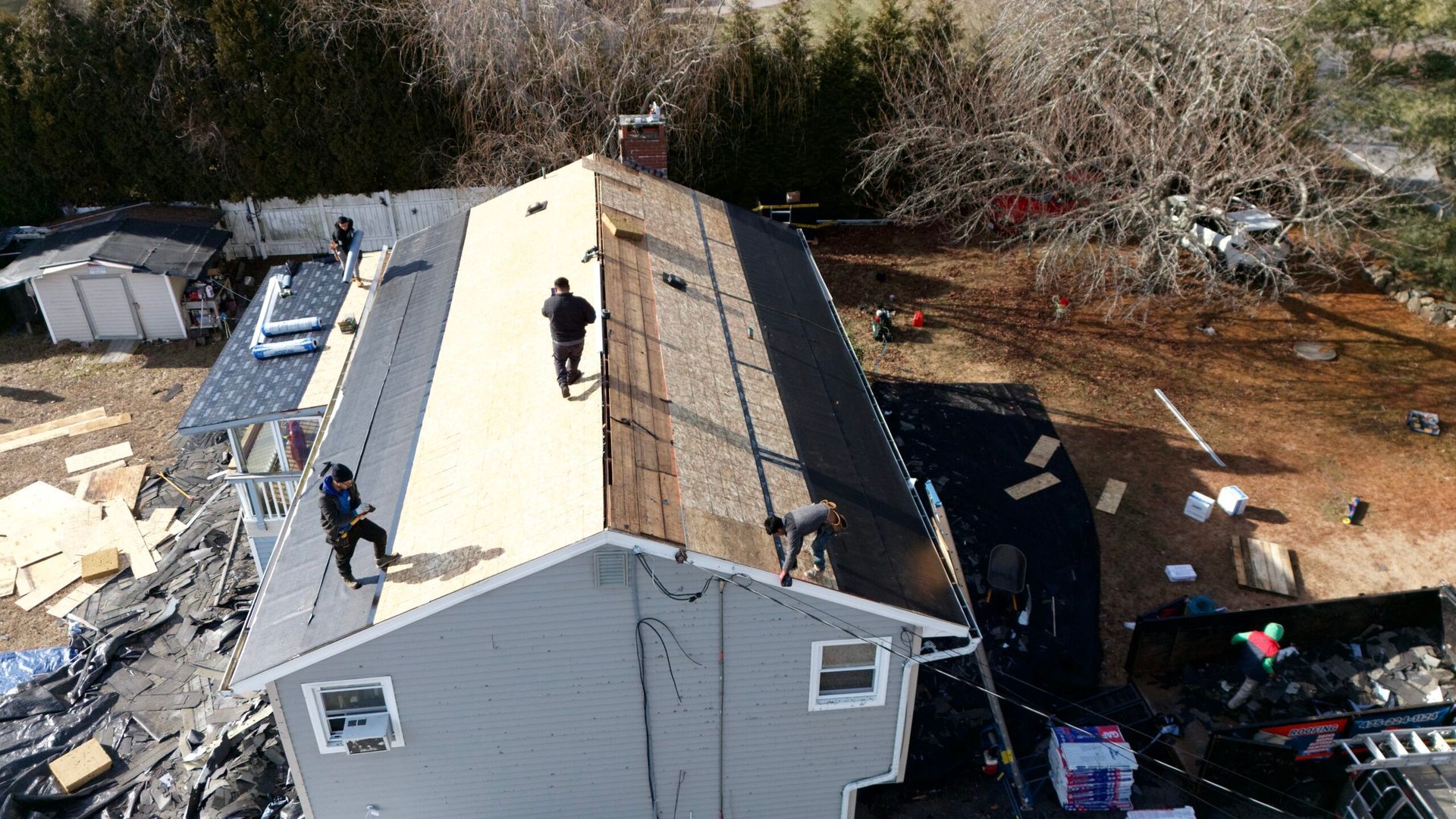 Aerial view of a roof under repair with workers and scattered roofing materials рядом a wooded area