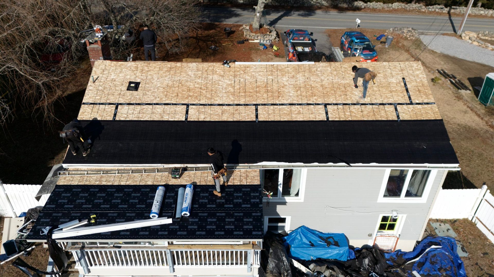 Aerial view of a house with a new roof under construction, workers on the roof and materials stacked nearby.
