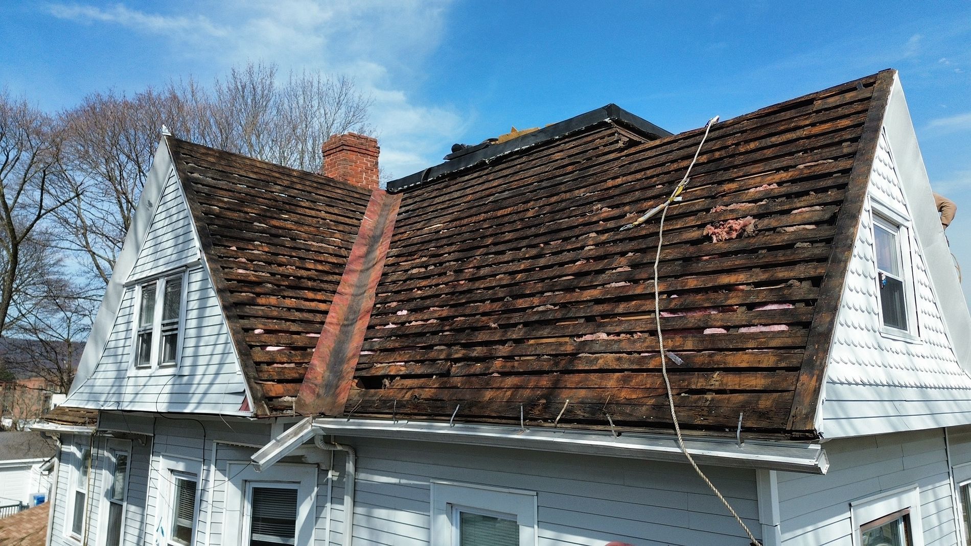 Snow-dusted house roof with peeling shingles and white siding under a clear blue sky