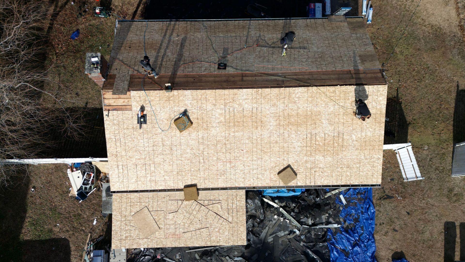 Aerial view of a house roof under construction with exposed plywood and surrounding yard equipment.
