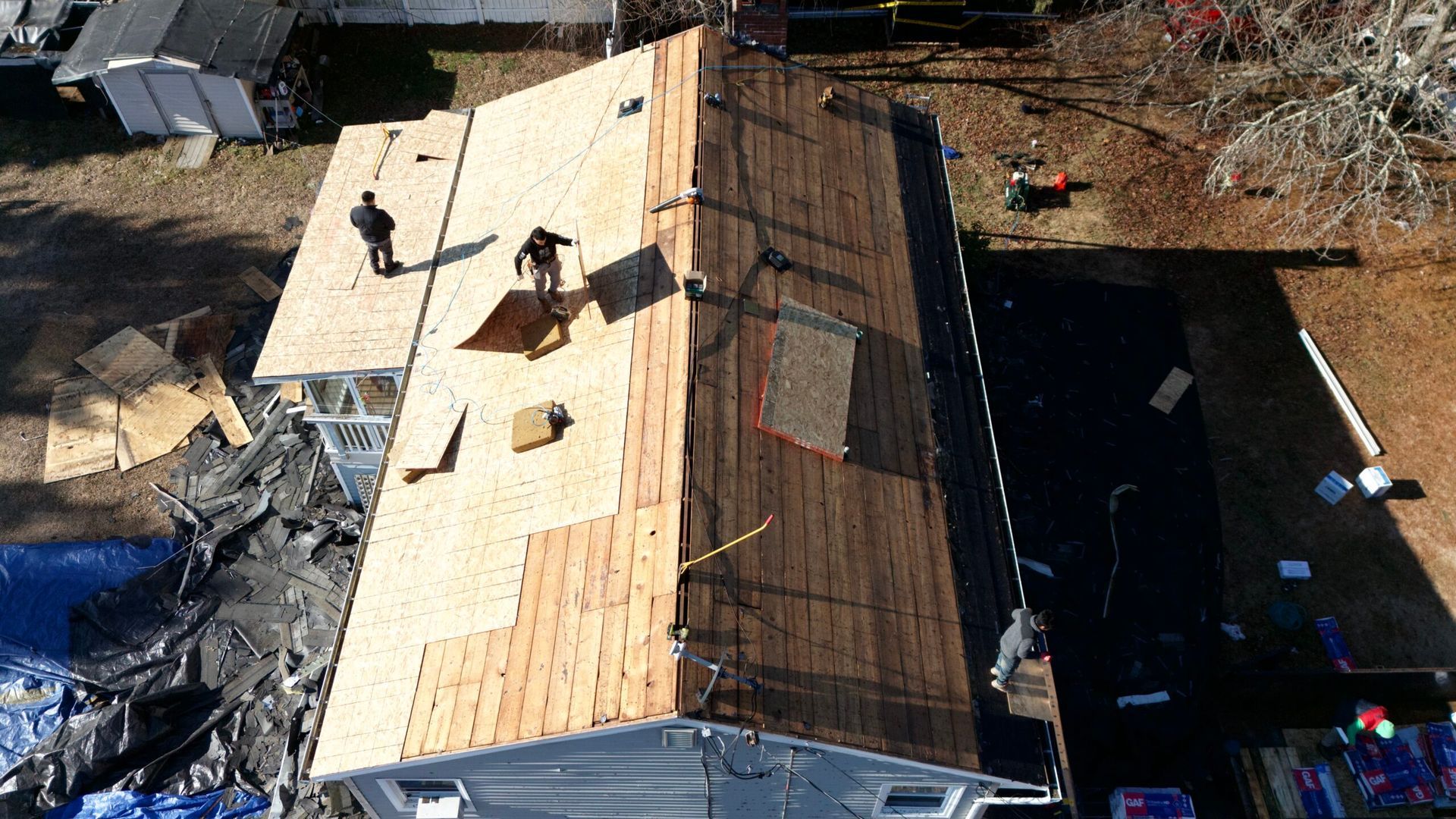 Aerial view of roofers installing tan shingles on a steep house roof beside a dark roof and yard.