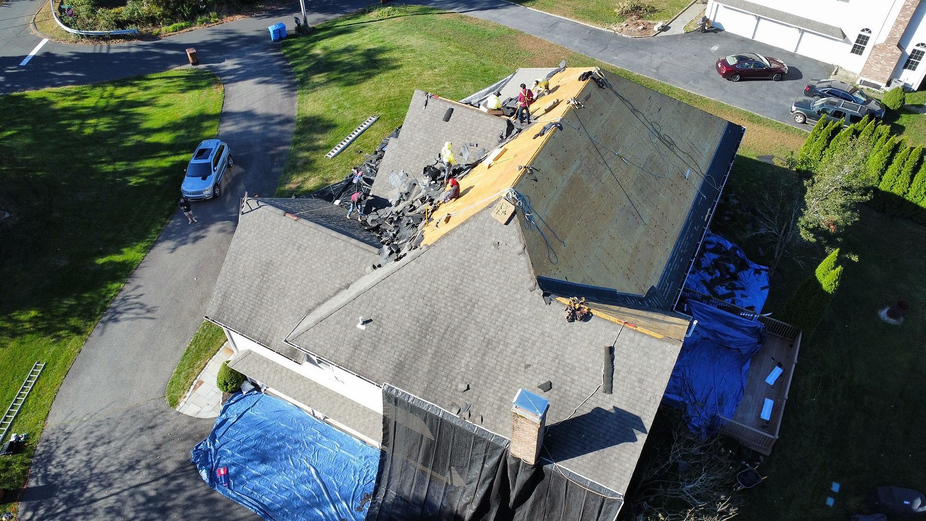 Aerial view of a house roof with workers repairing damaged shingles and blue tarp sections.