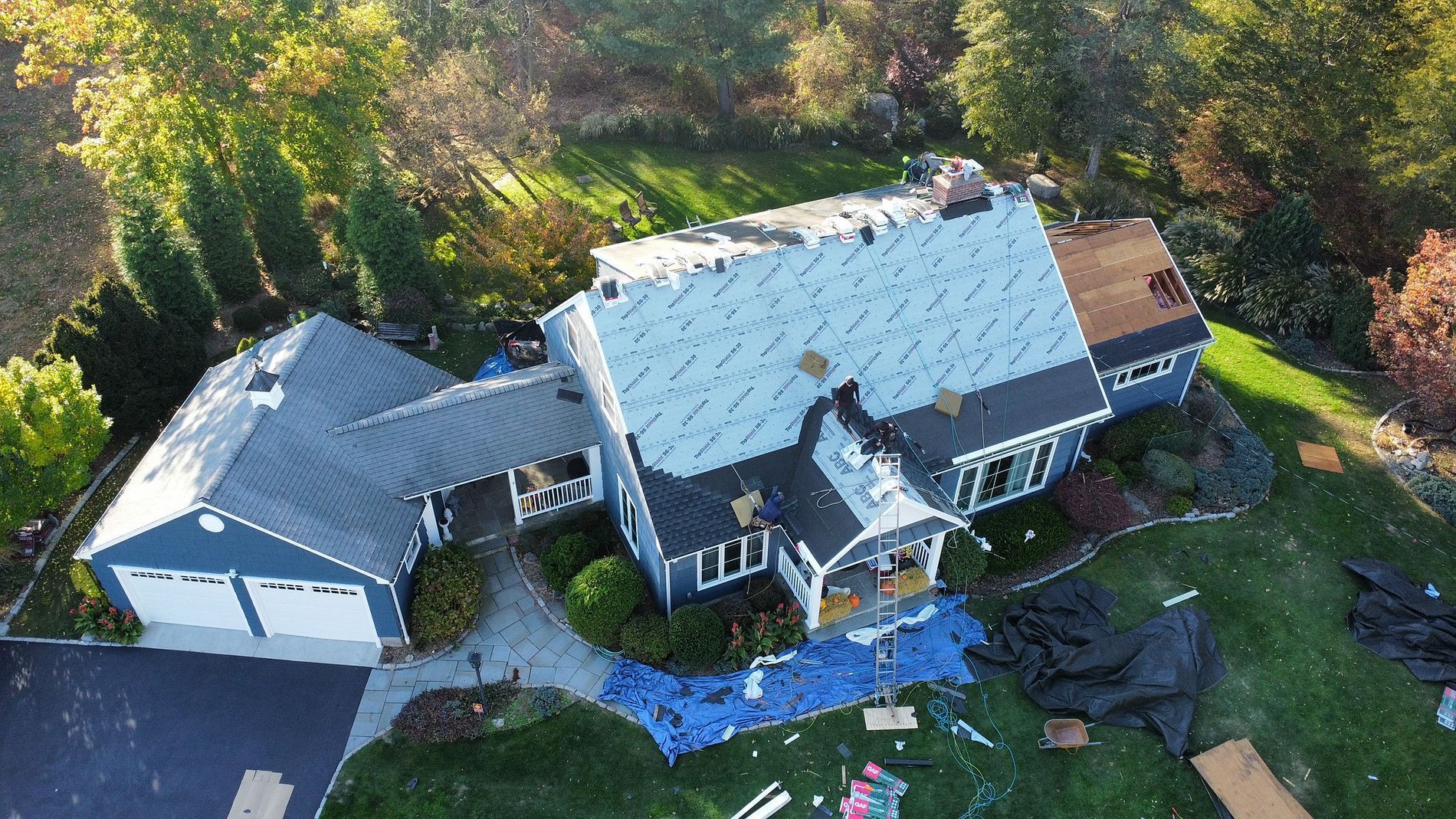 Aerial view of a large gray house with a detached garage, surrounded by trees and a landscaped yard.