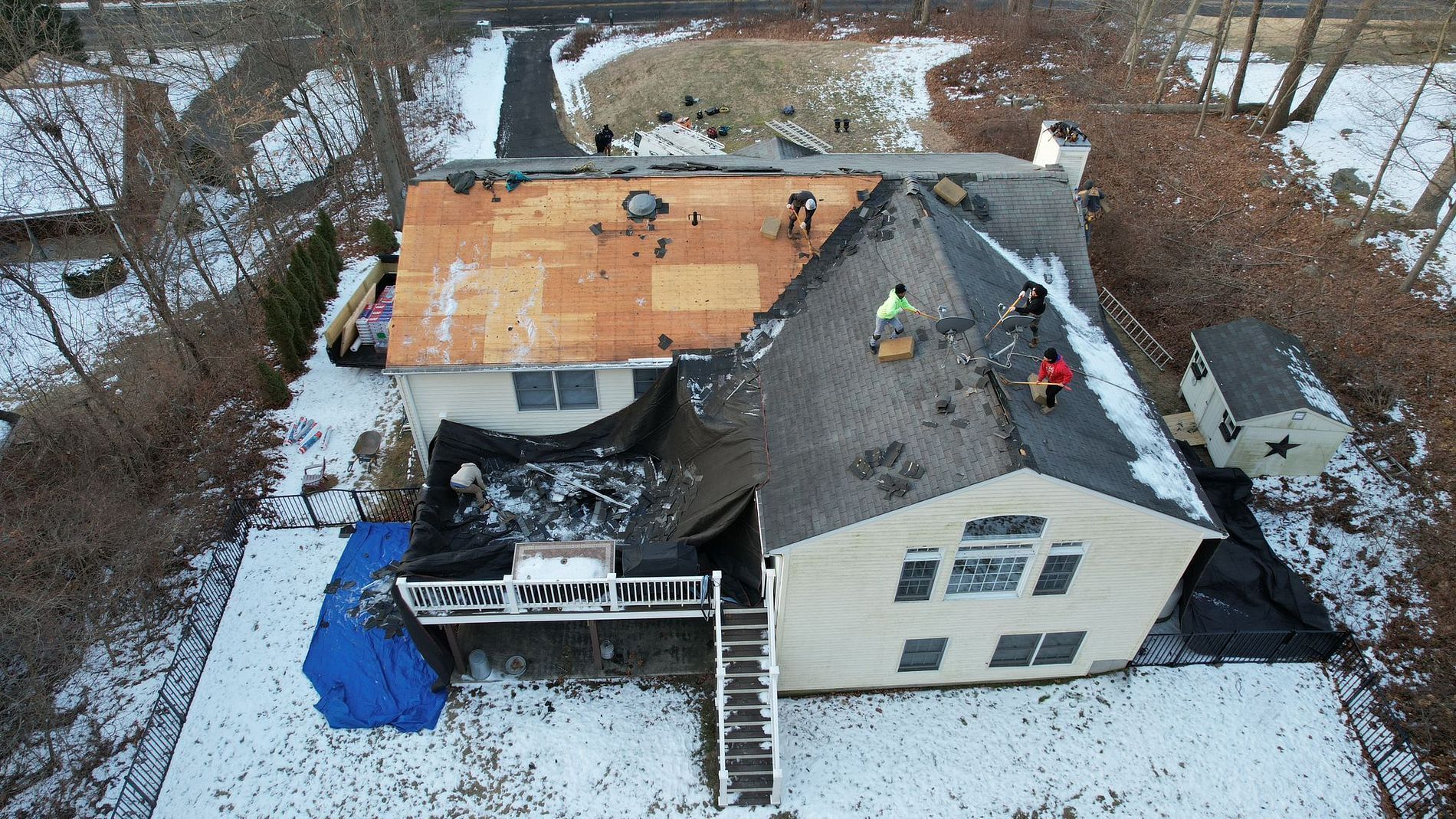 Aerial view of a house with a roof being repaired, workers on top, and snow-covered ground вокруг