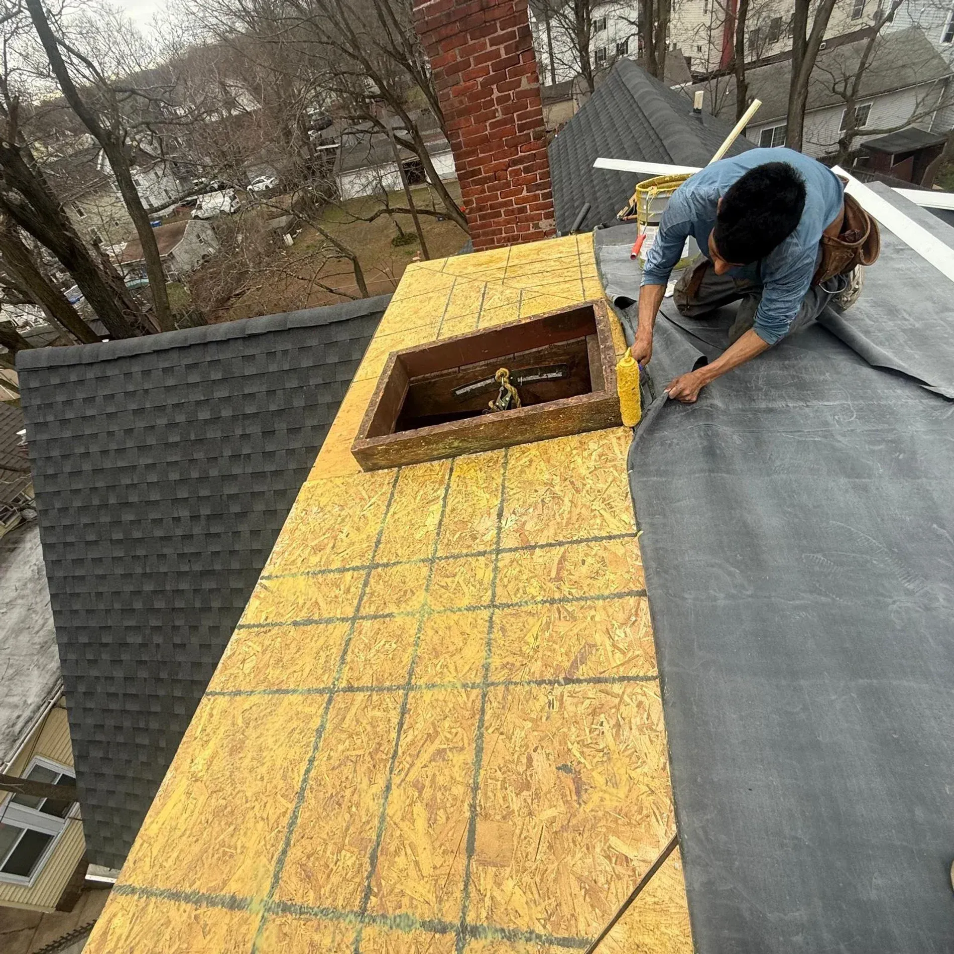 Worker installing roofing felt beside a brick chimney on a sloped roof