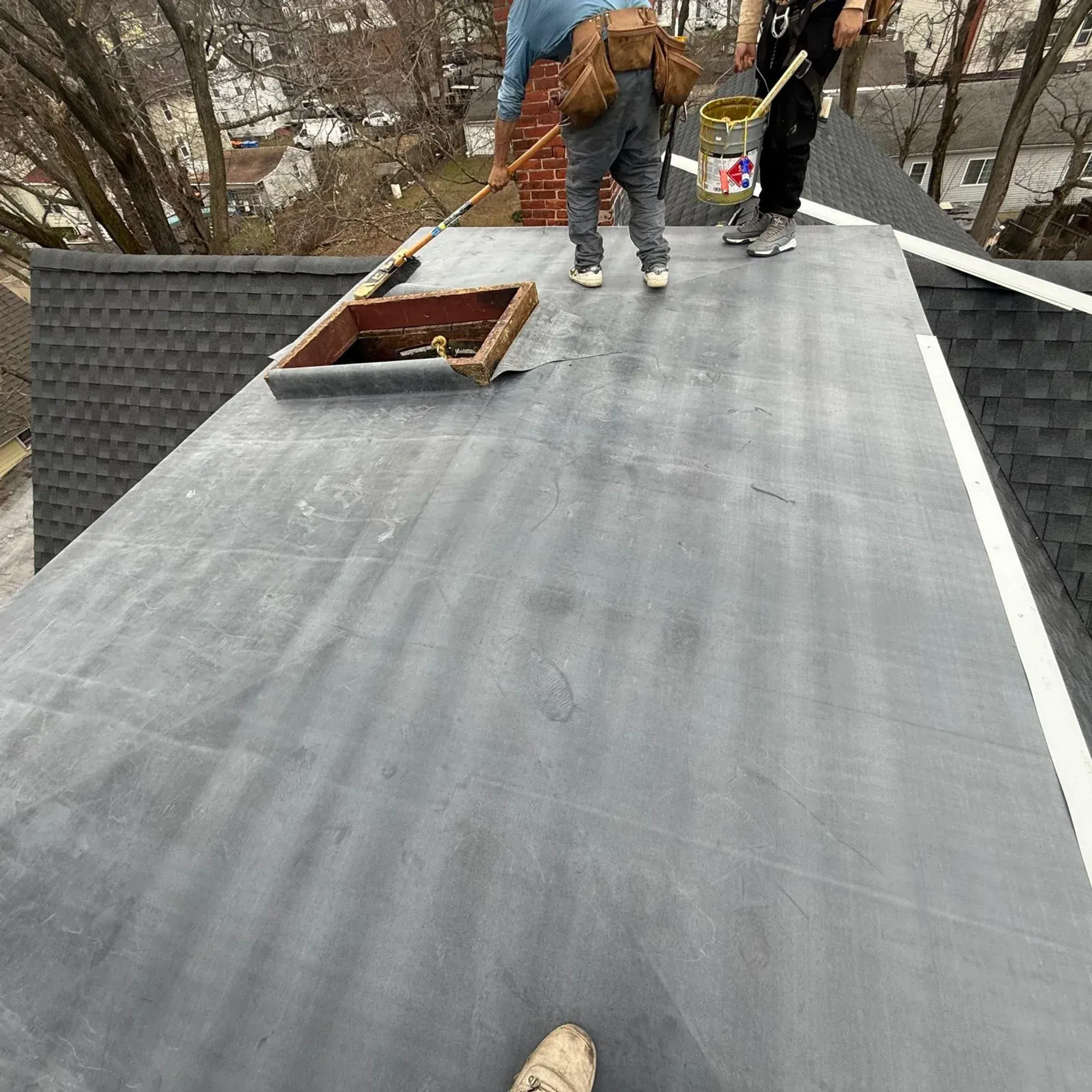 Workers on a gray roof installing roofing material near a chimney opening.