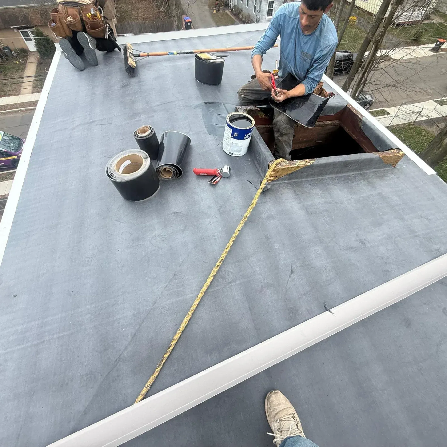 Worker repairing a flat roof near an open hatch, with tools and a safety rope visible.