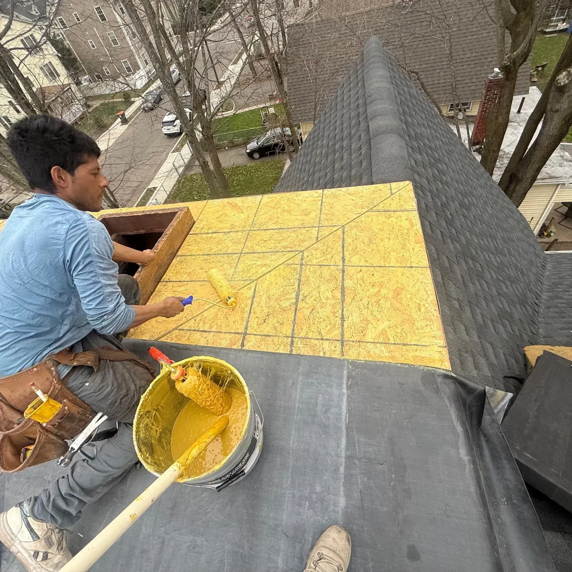 Person roofing on a steep roof, spreading yellow sealant near a bucket and trowel