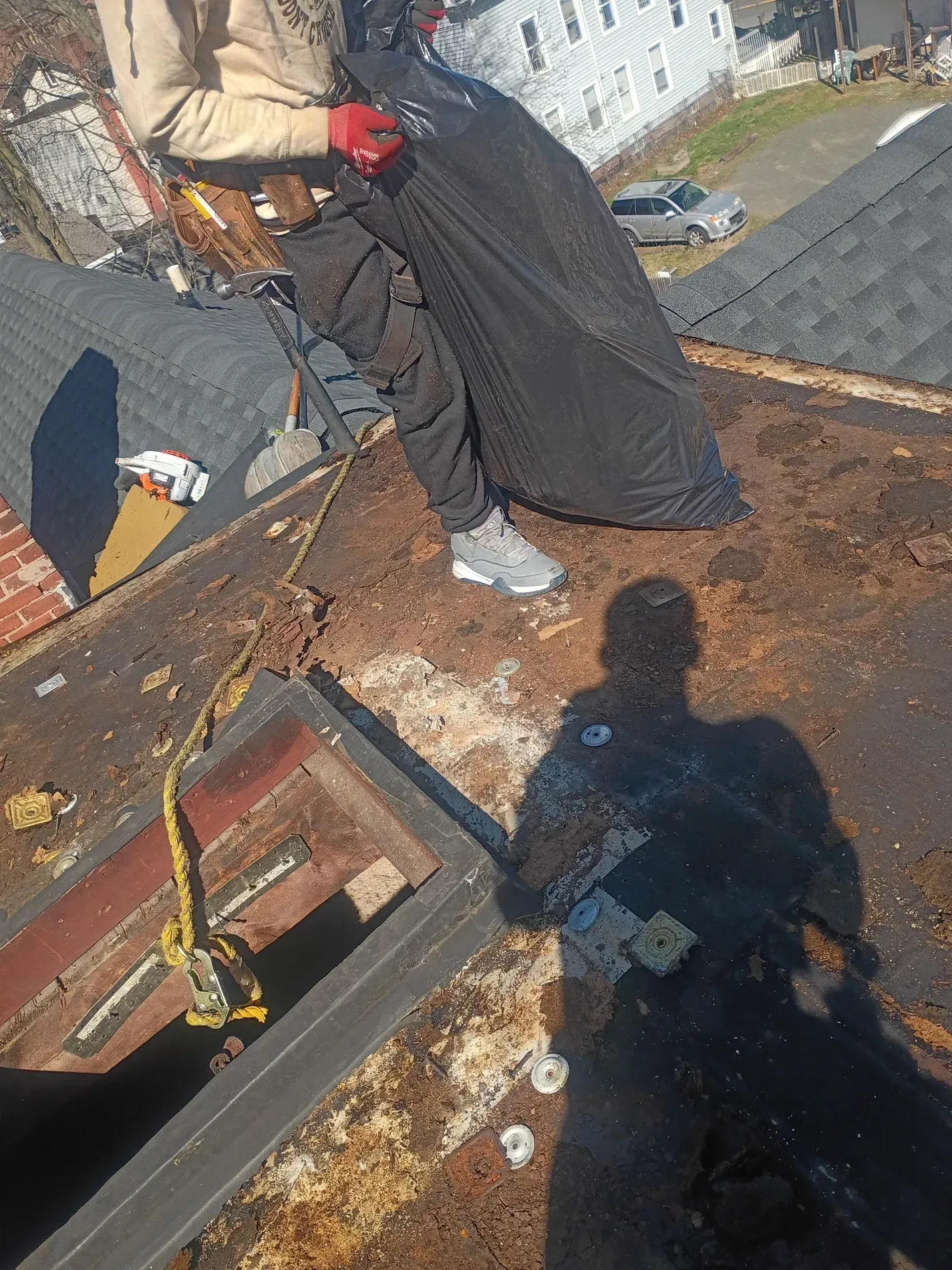 Worker on a steep rooftop near a chimney, securing materials beside a black safety tarp.