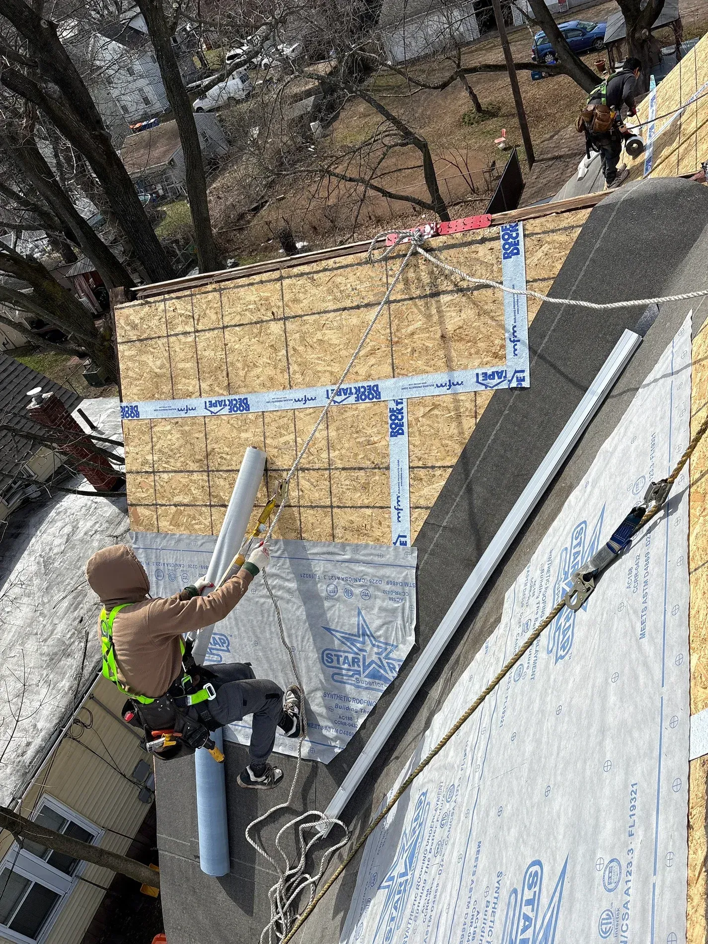 Worker installing roof sheathing on a steep roof, with blue tape and plywood panels visible.