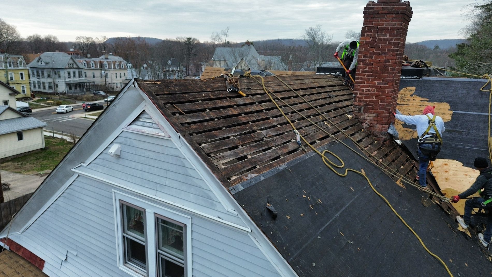 Workers repairing a roof with exposed plywood and a brick chimney in a residential neighborhood