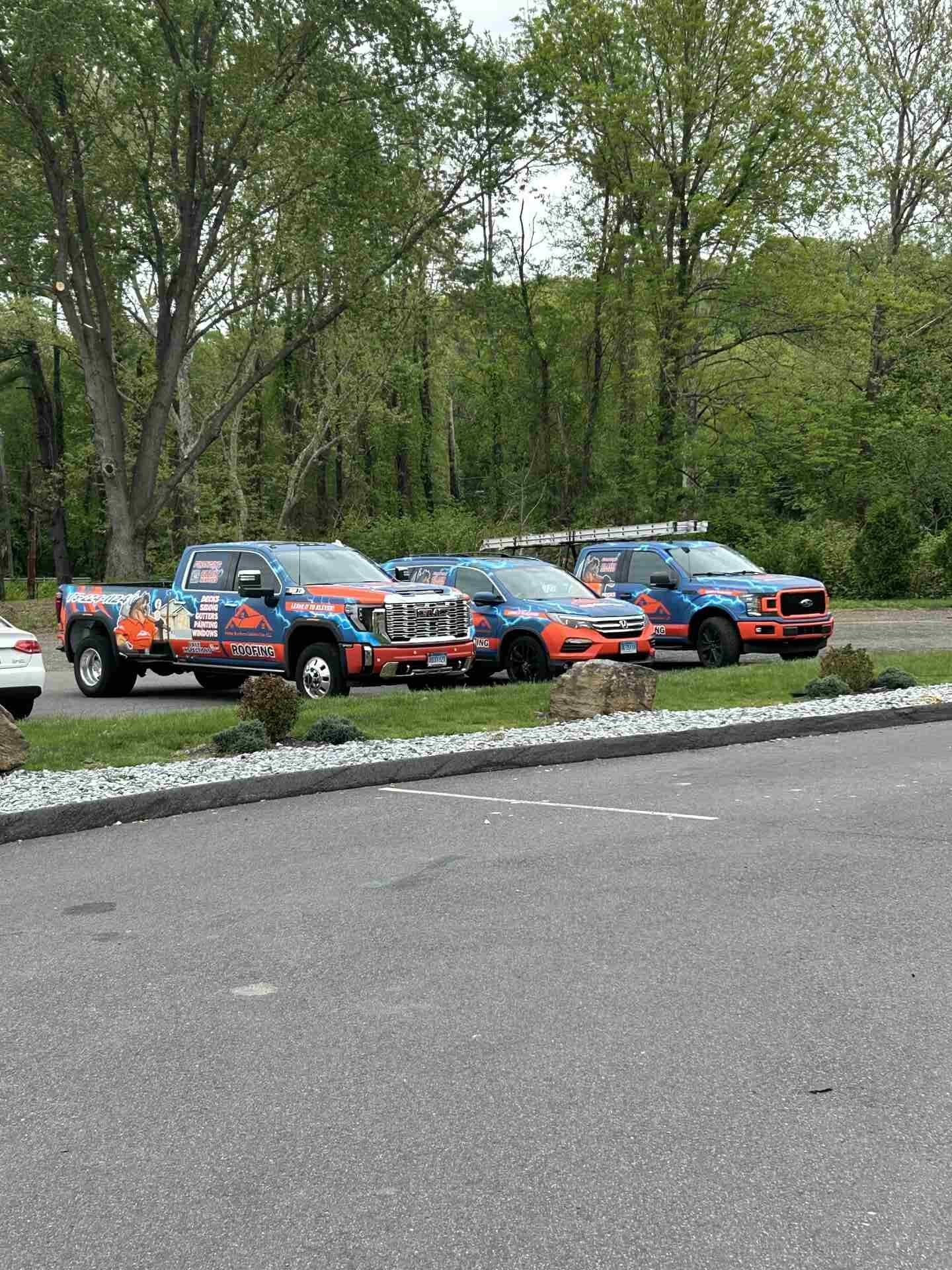 Three rally cars in blue and orange parked on a gravel roadside beside trees.