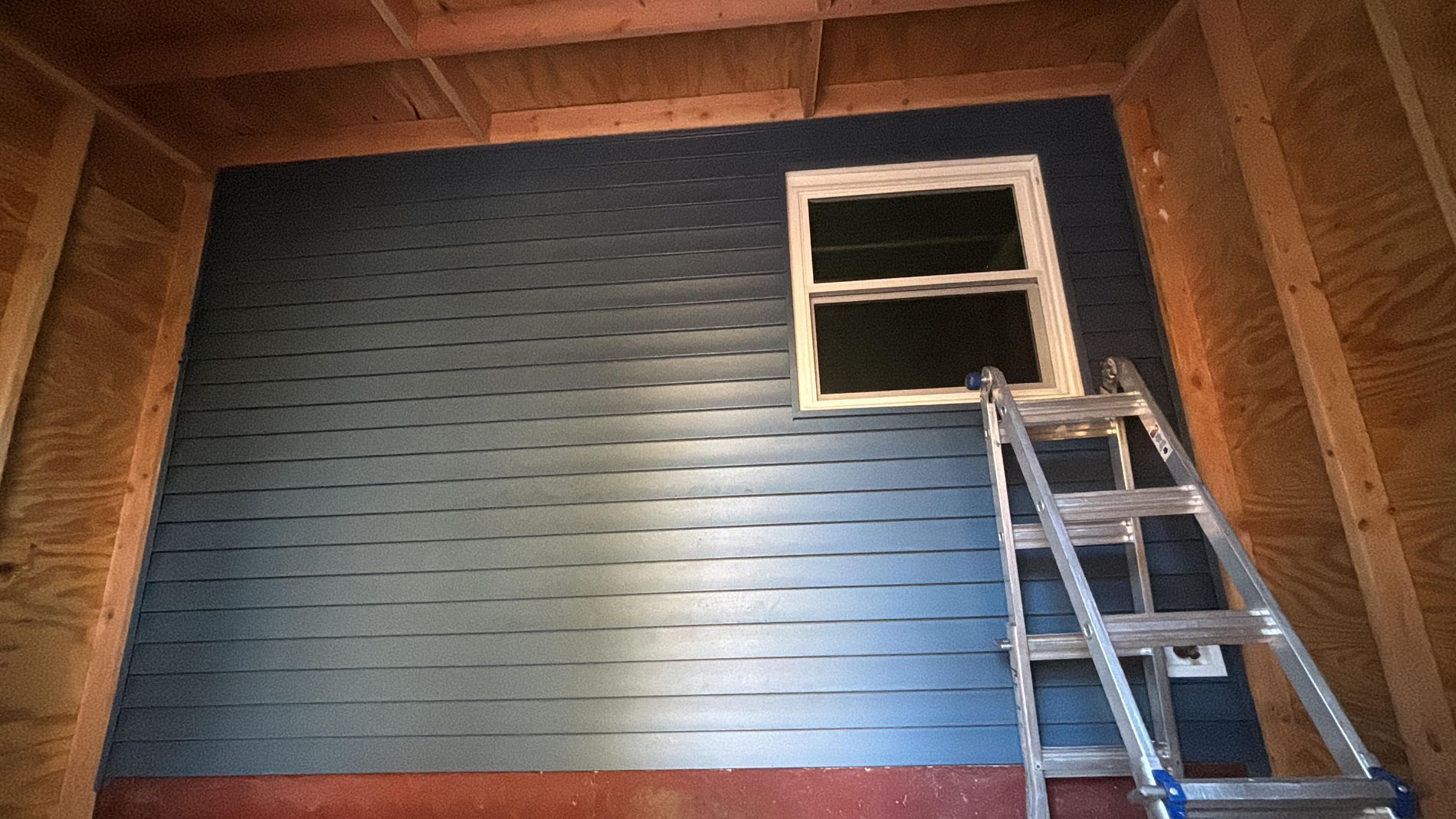 Blue wooden wall with a small window and a ladder leaning against it in an attic-like room
