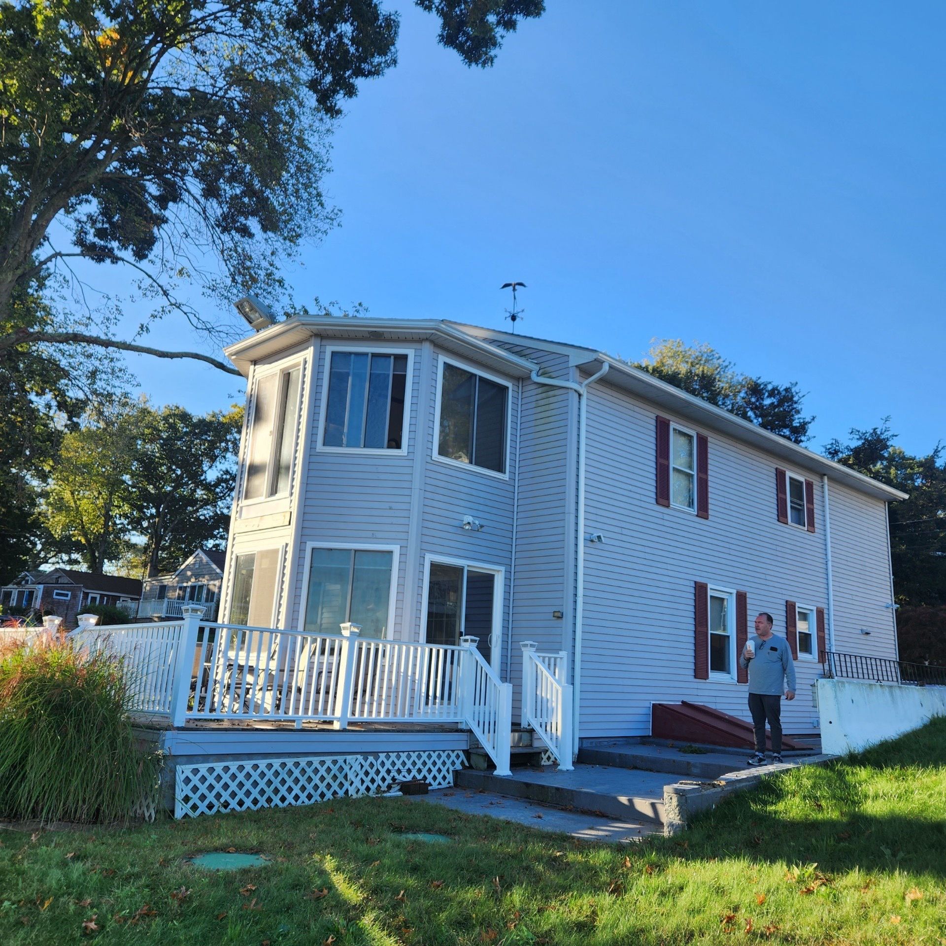 White two-story house with blue-trimmed porch and a person standing by the side steps in a sunny yard