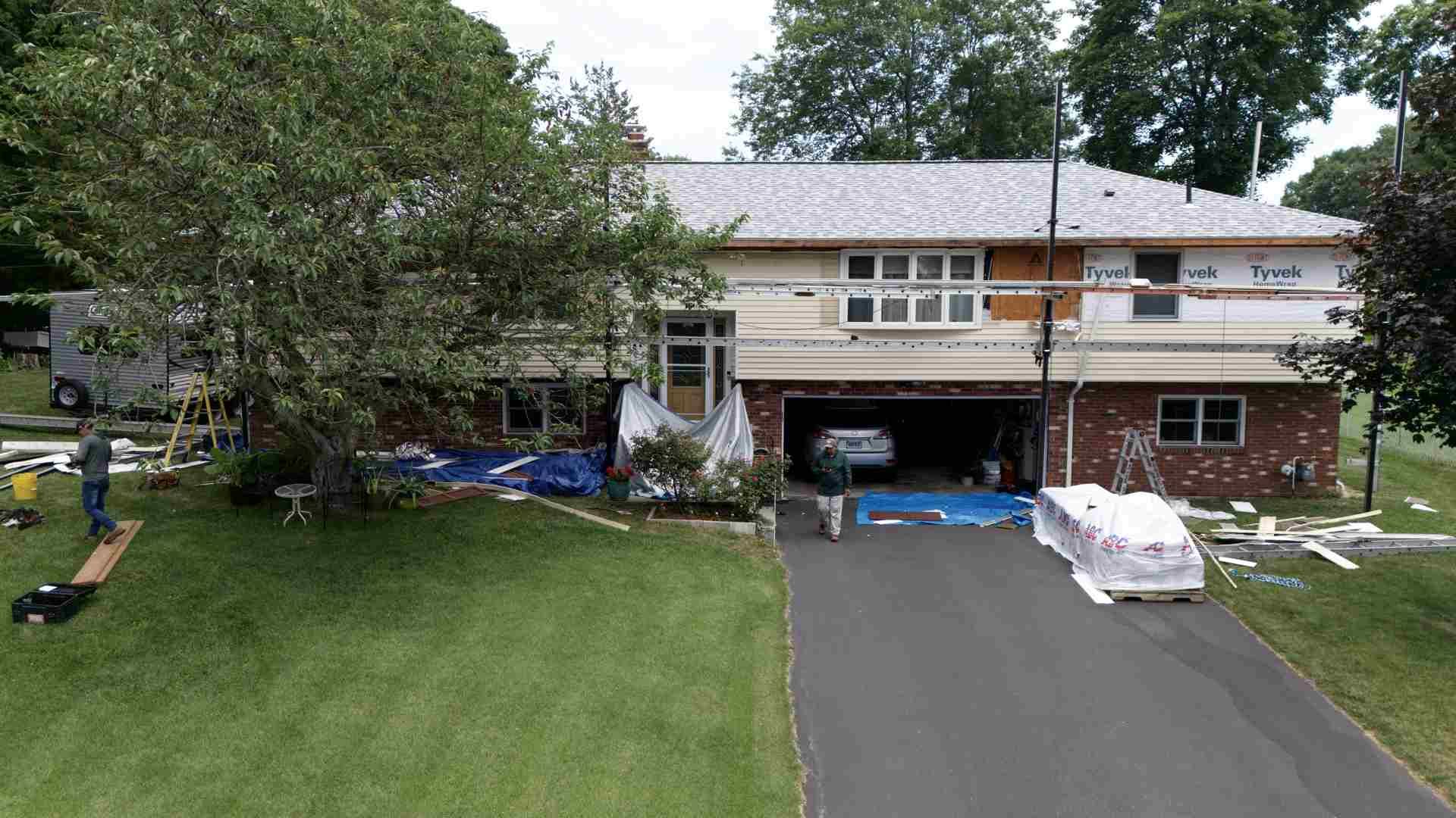 Suburban house with driveway, garage, and large green front lawn; construction materials cover part of the facade.