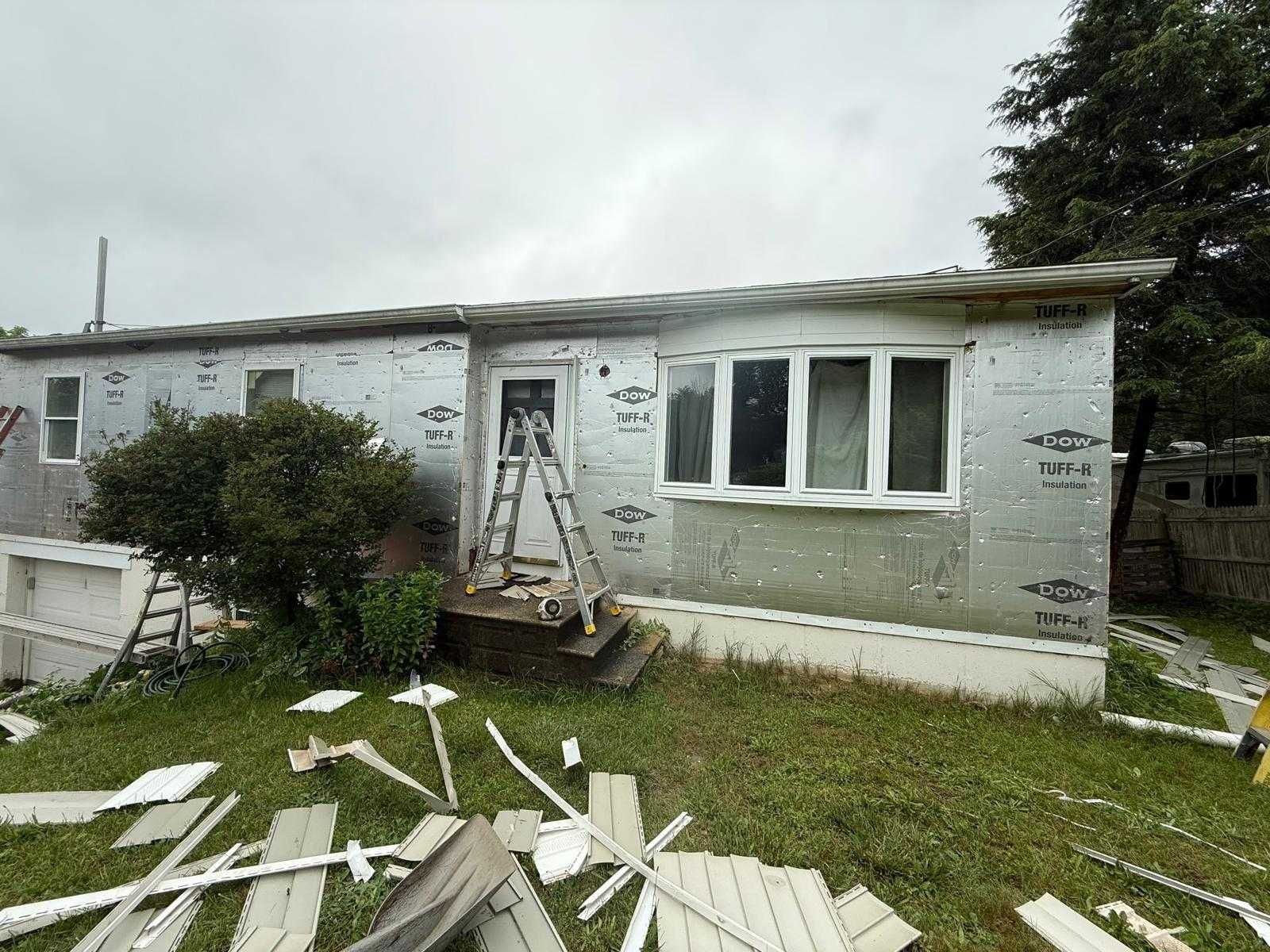 Damaged white house exterior with a ladder, broken siding, and debris scattered on the lawn