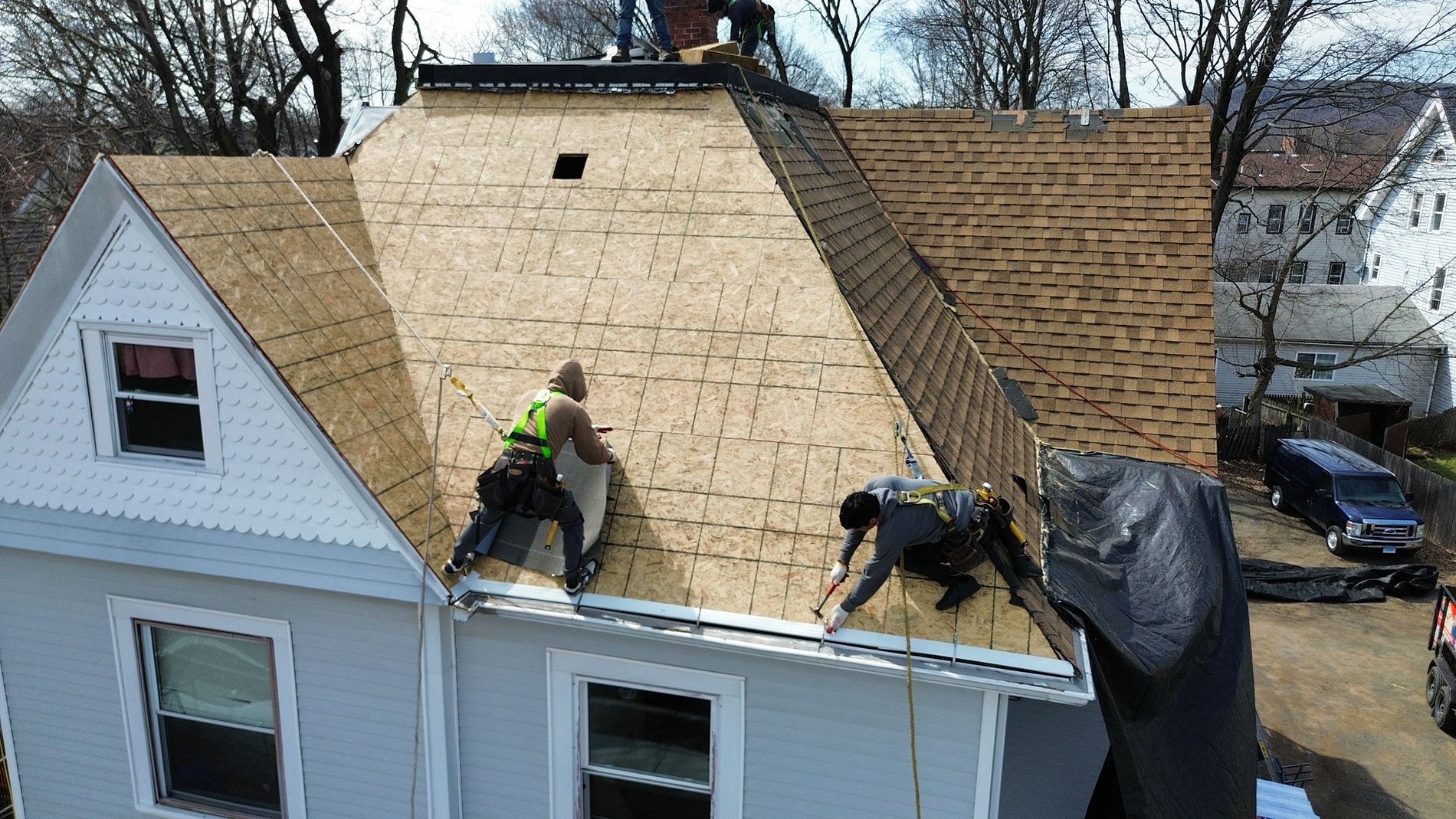 Roofers installing plywood on a house roof during repairs, with shingles removed and a tarp on one side.