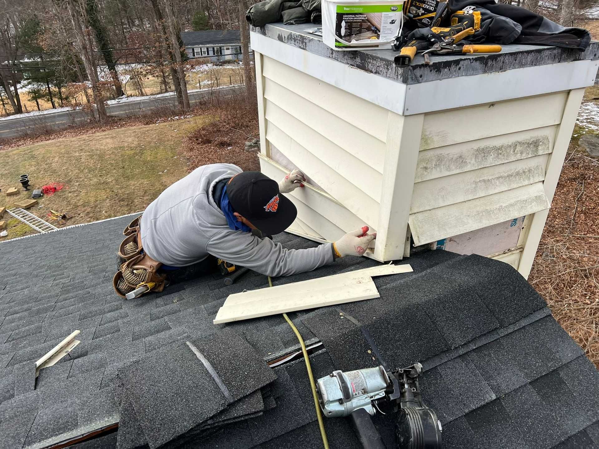 Worker repairing shingles around a white chimney on a gray roof