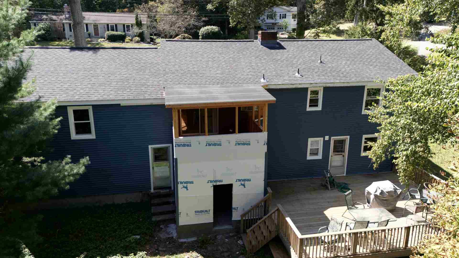 Blue house with a gray roof, front porch, and wooden deck surrounded by trees.