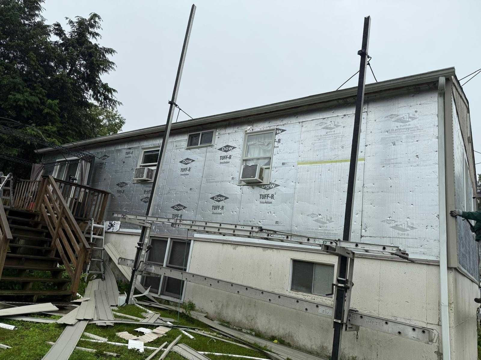 Damaged white house exterior with leaning poles, peeling siding, and a small stairway in a grassy yard