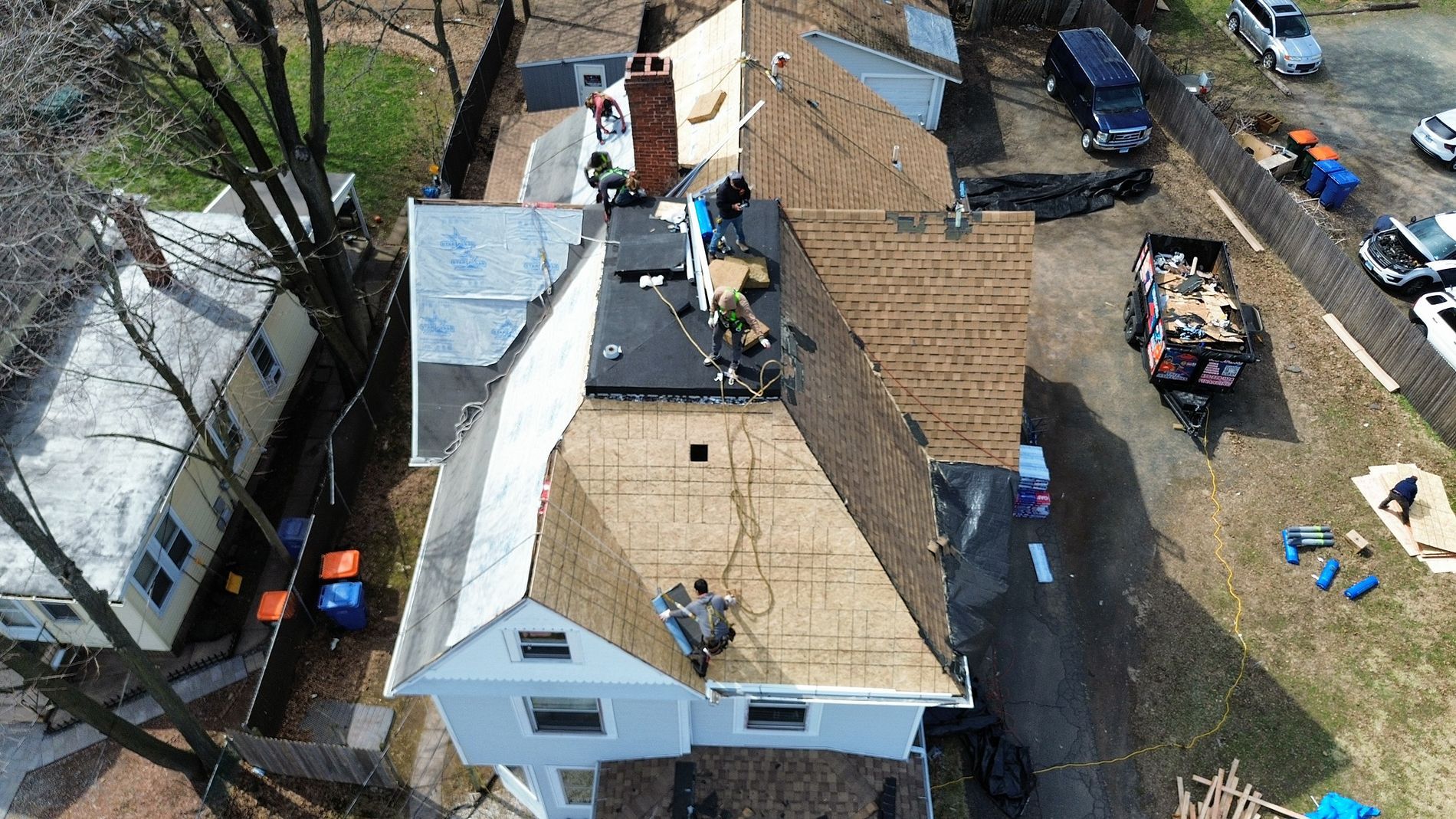 Aerial view of workers repairing a house roof beside parked vehicles and construction materials.