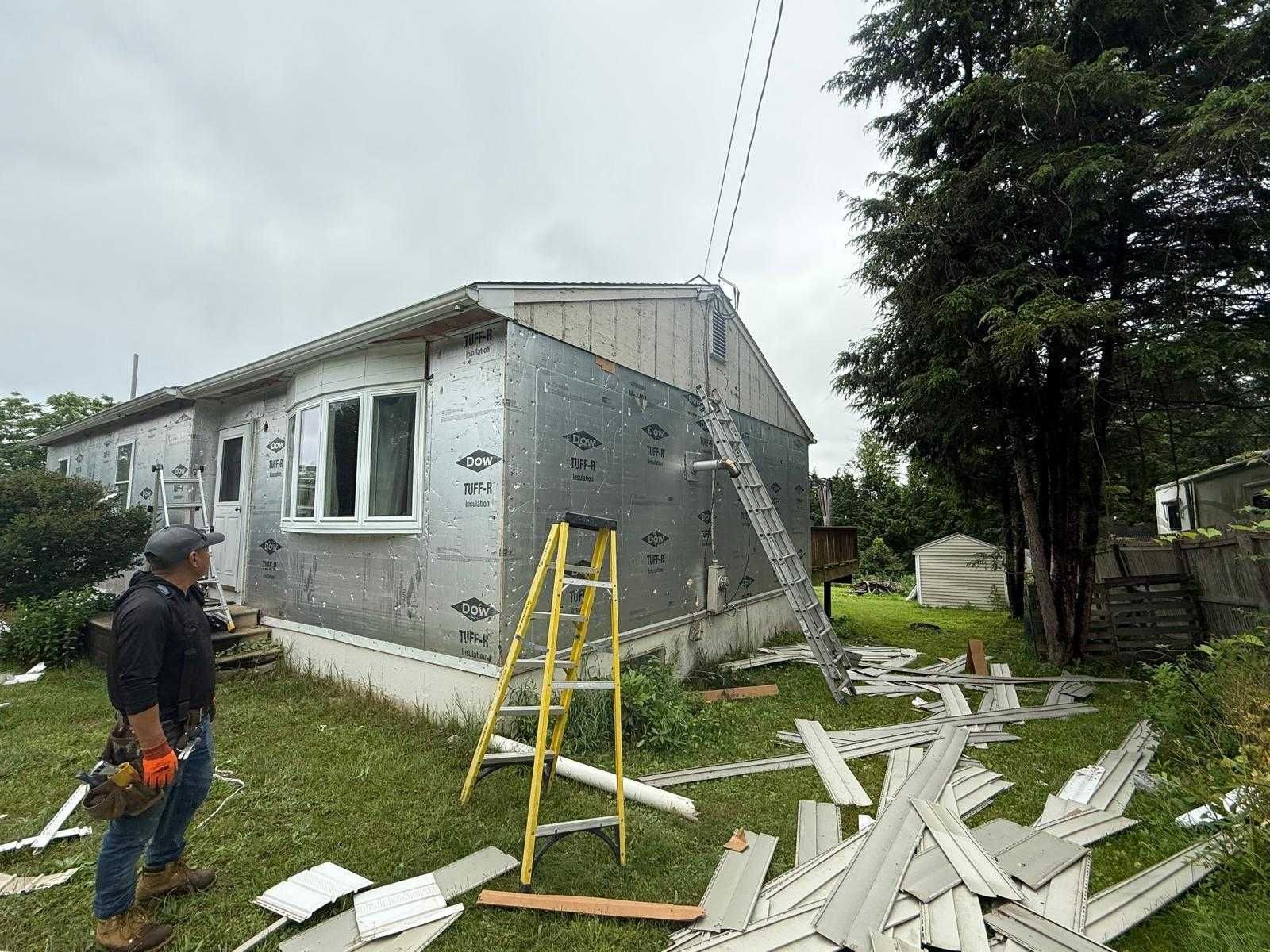 Workers repairing a damaged house exterior with ladders, debris, and siding scattered on the lawn