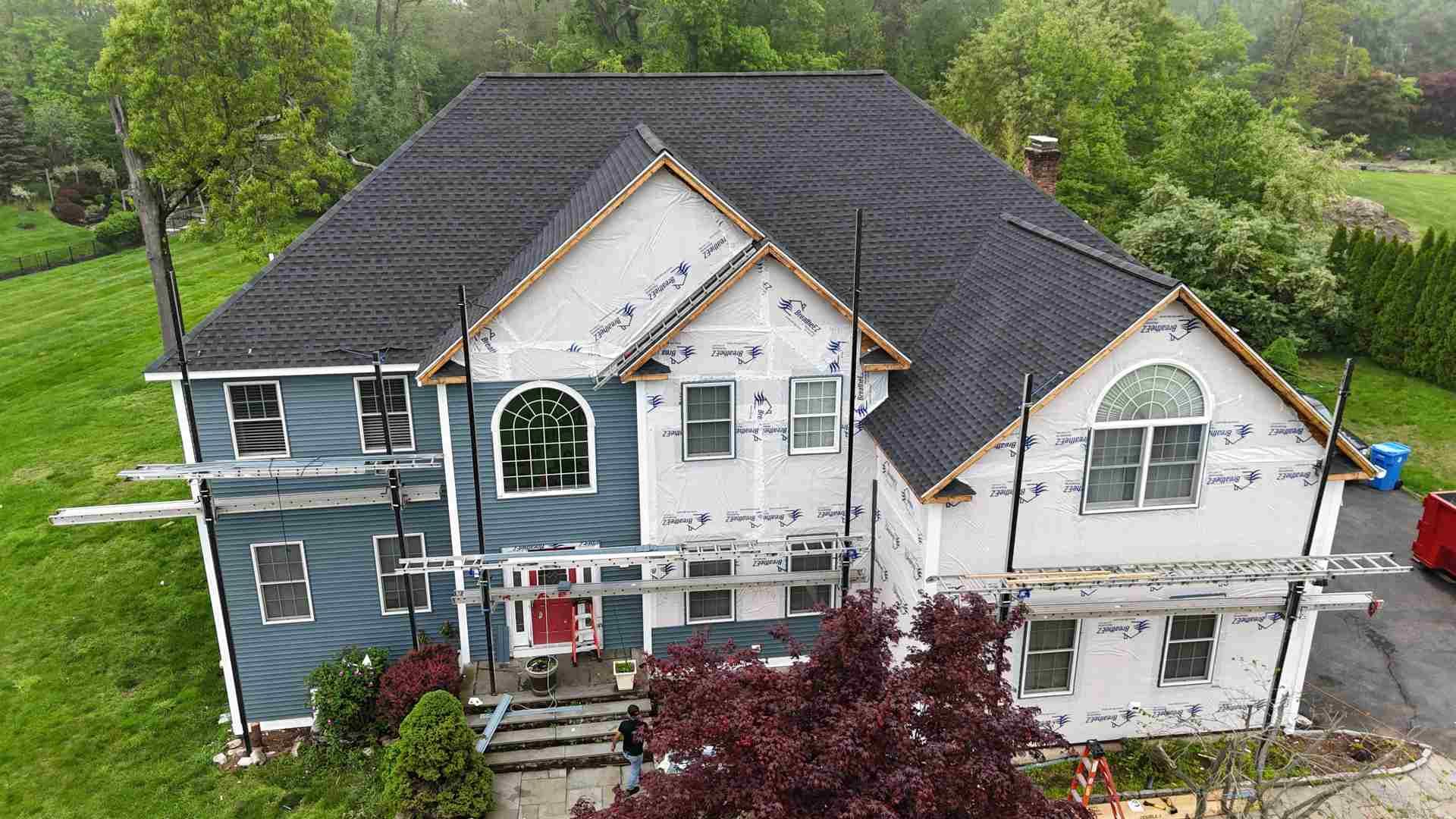 Large two-story house with gray, white, and blue siding and a dark shingled roof, surrounded by trees and lawn.