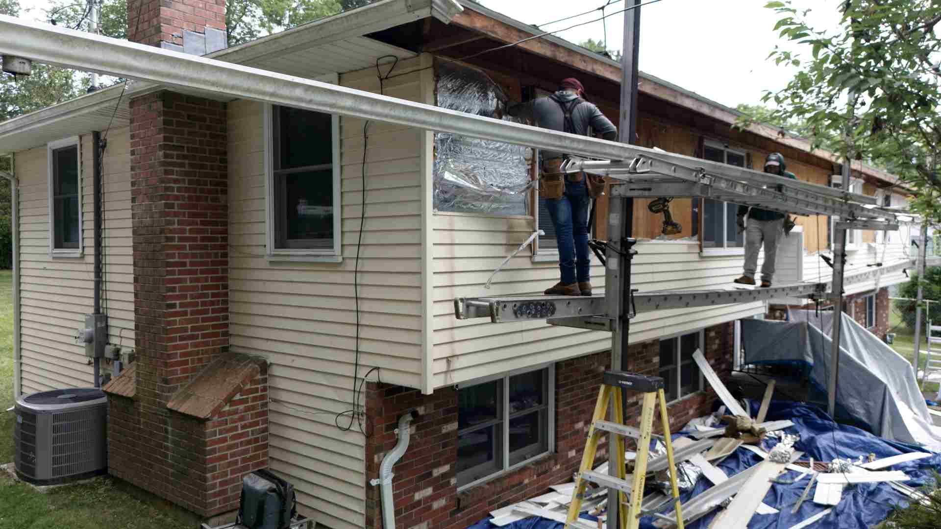 Workers on a balcony installing siding and railing on a house exterior, with construction debris below.