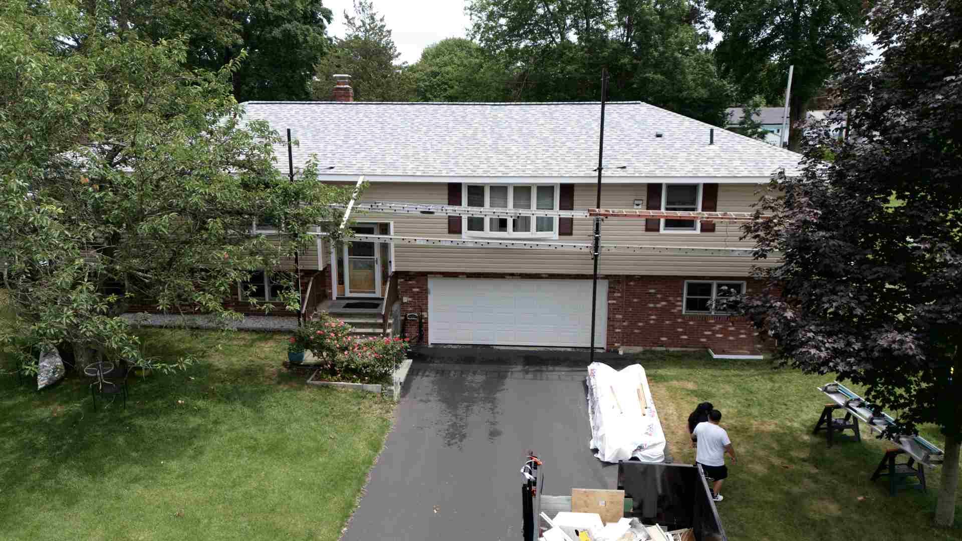 Aerial view of a suburban house with a driveway, two people, and trees surrounding the yard.