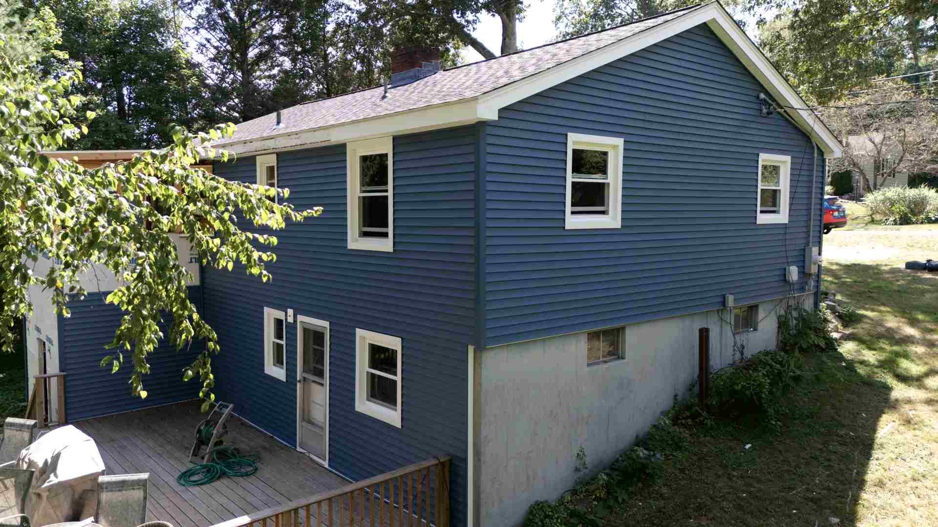 Blue two-story house with white trim and a raised lower level beside trees and a fenced yard