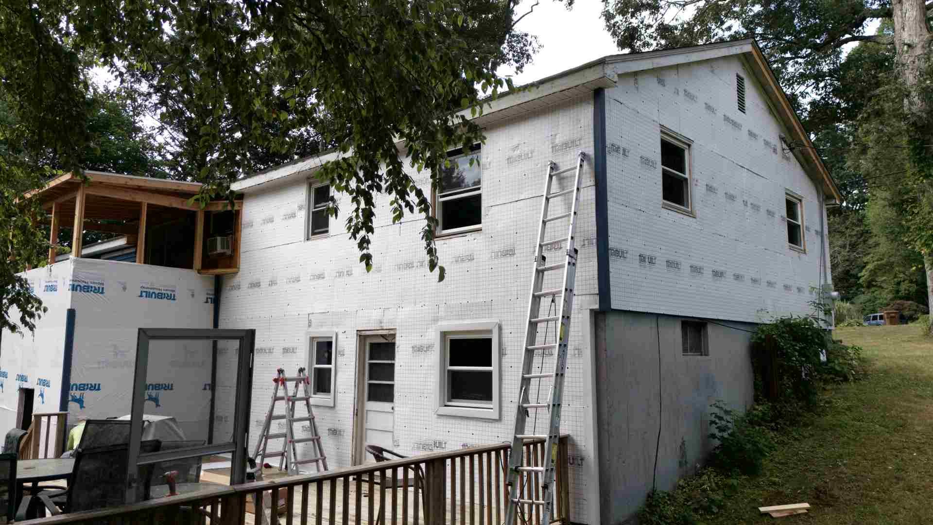 Partially renovated white house with exposed siding, ladders, and scaffolding under trees