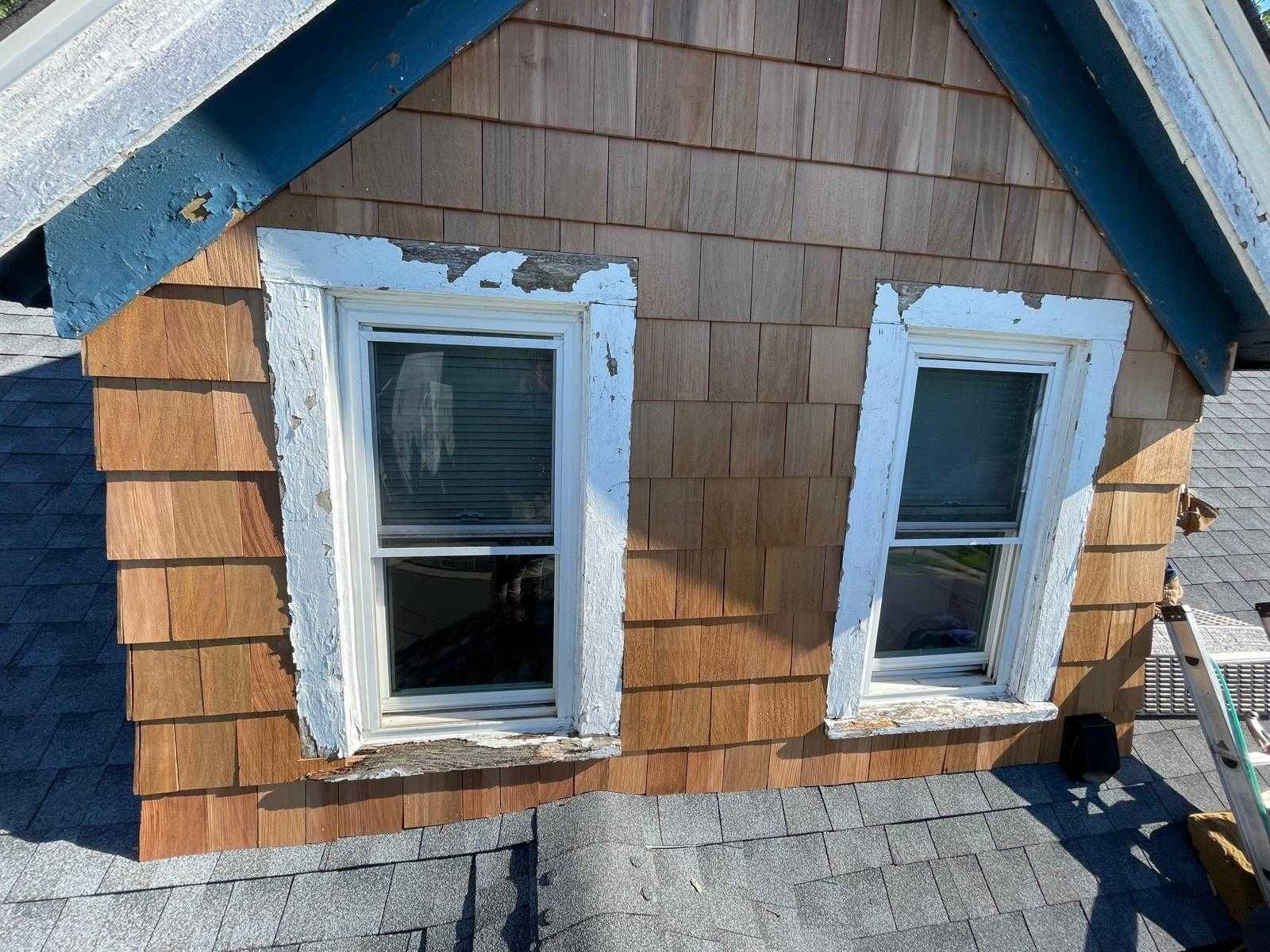 Weathered house exterior with two white-trimmed windows, tan shingles, and blue trim above a roofline