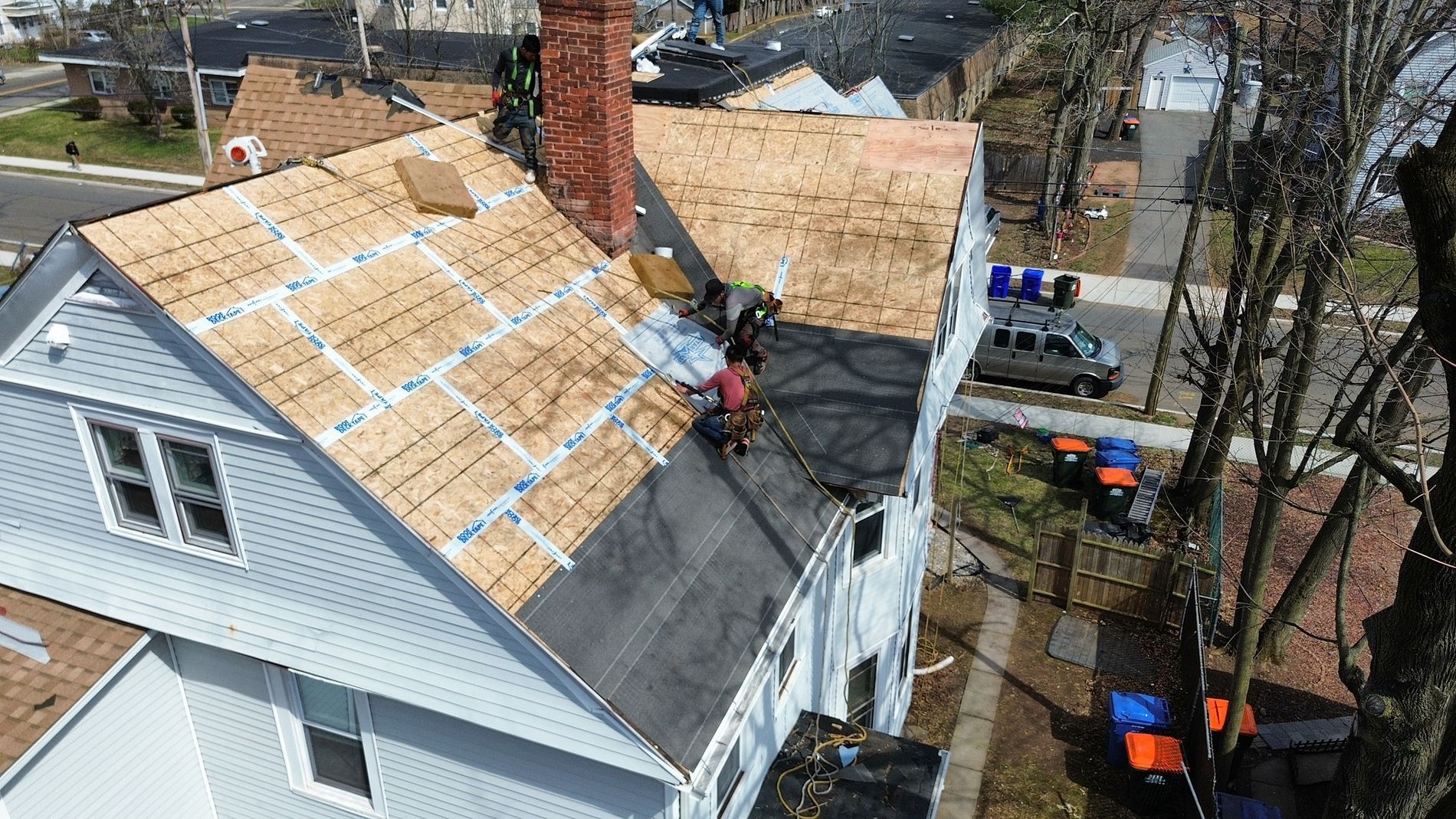 Roofers replacing a steep white house roof with plywood decking exposed and tools on a laddered roof.
