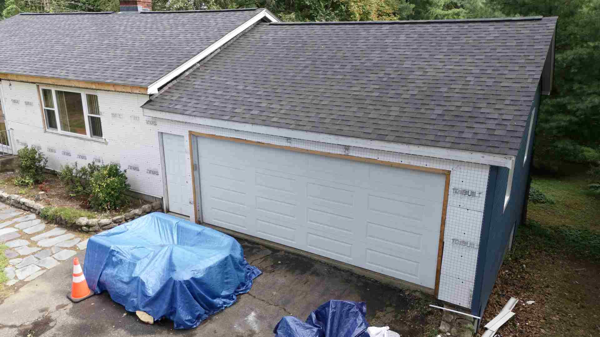 Single-story house with double garage, blue tarp-covered items in driveway, and roof work in progress