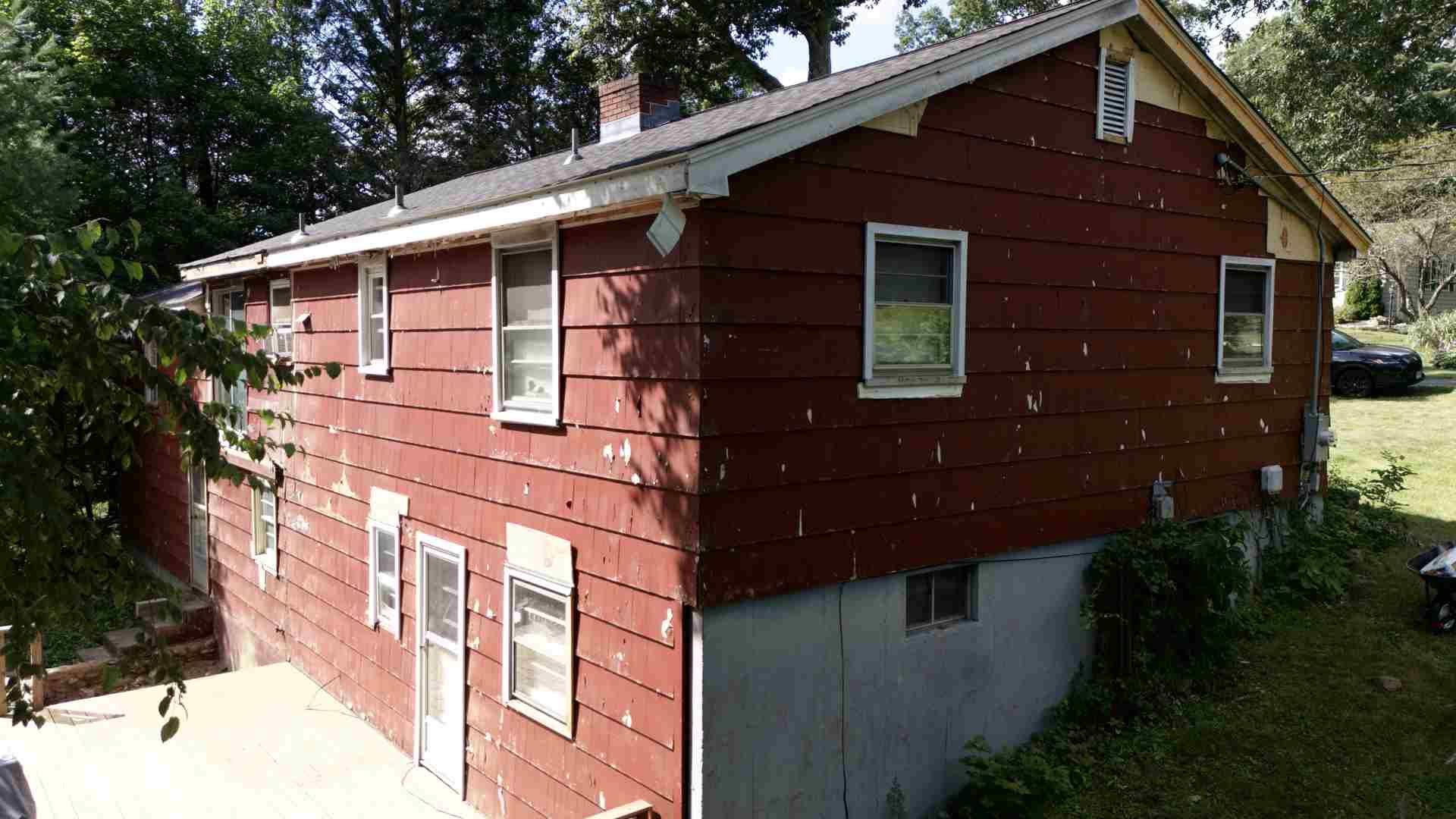 Red two-story house with white trim and a concrete foundation, viewed from the side in a wooded yard