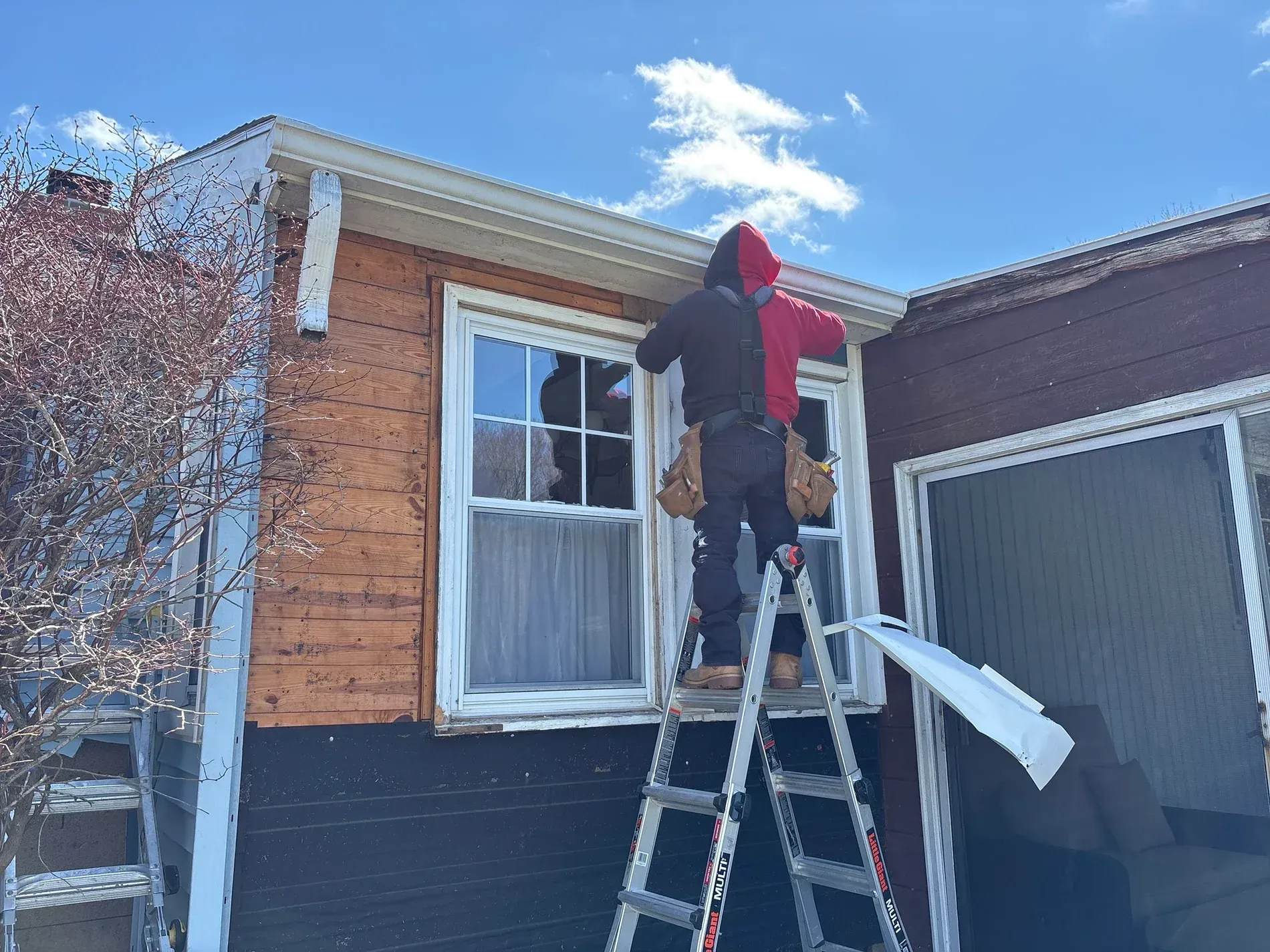 Worker on ladder installing siding above a window on a brick-and-black house