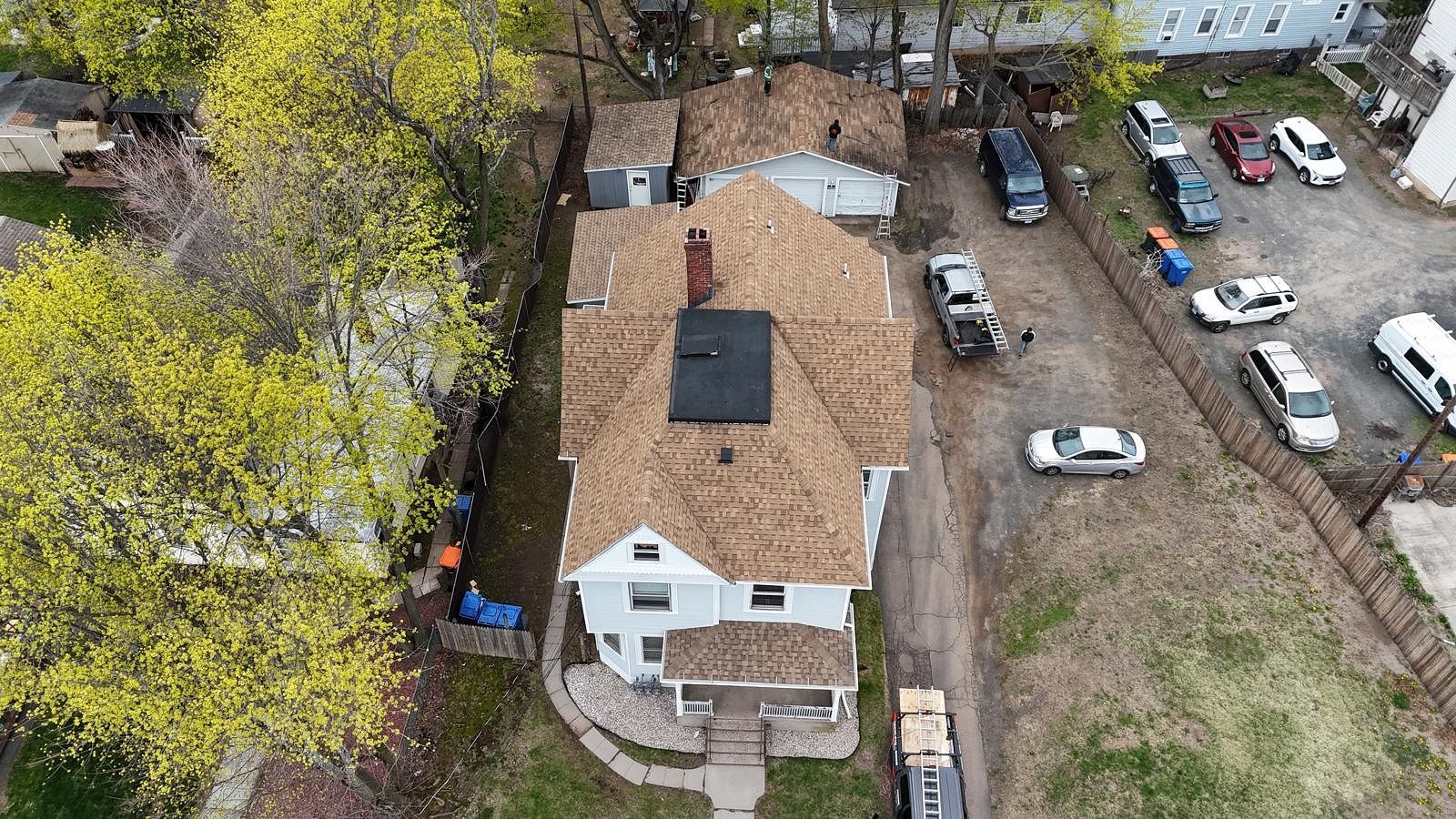 Aerial view of a white house with tan roof, surrounded by trees, yard, and parked cars.