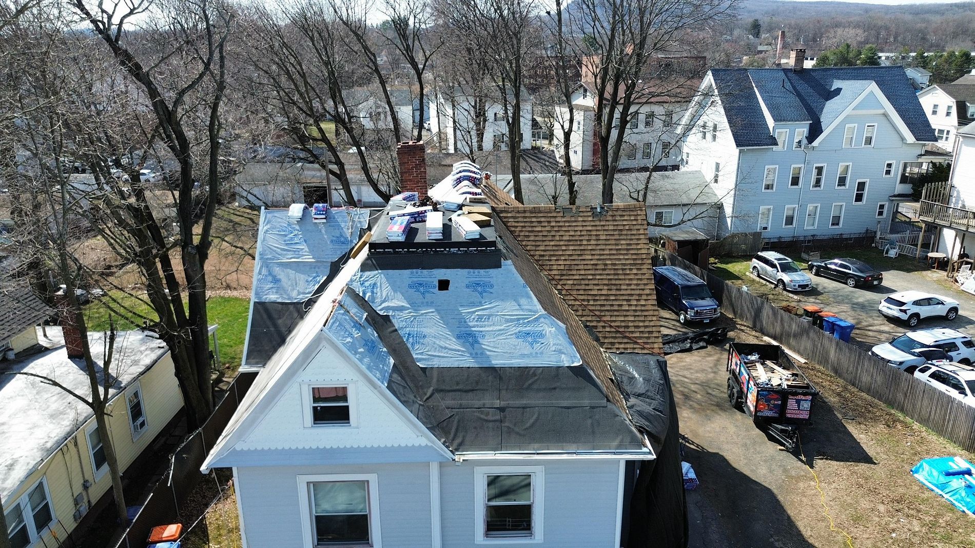Residential roof under repair with workers, exposed shingles, and neighboring houses in a snowy yard