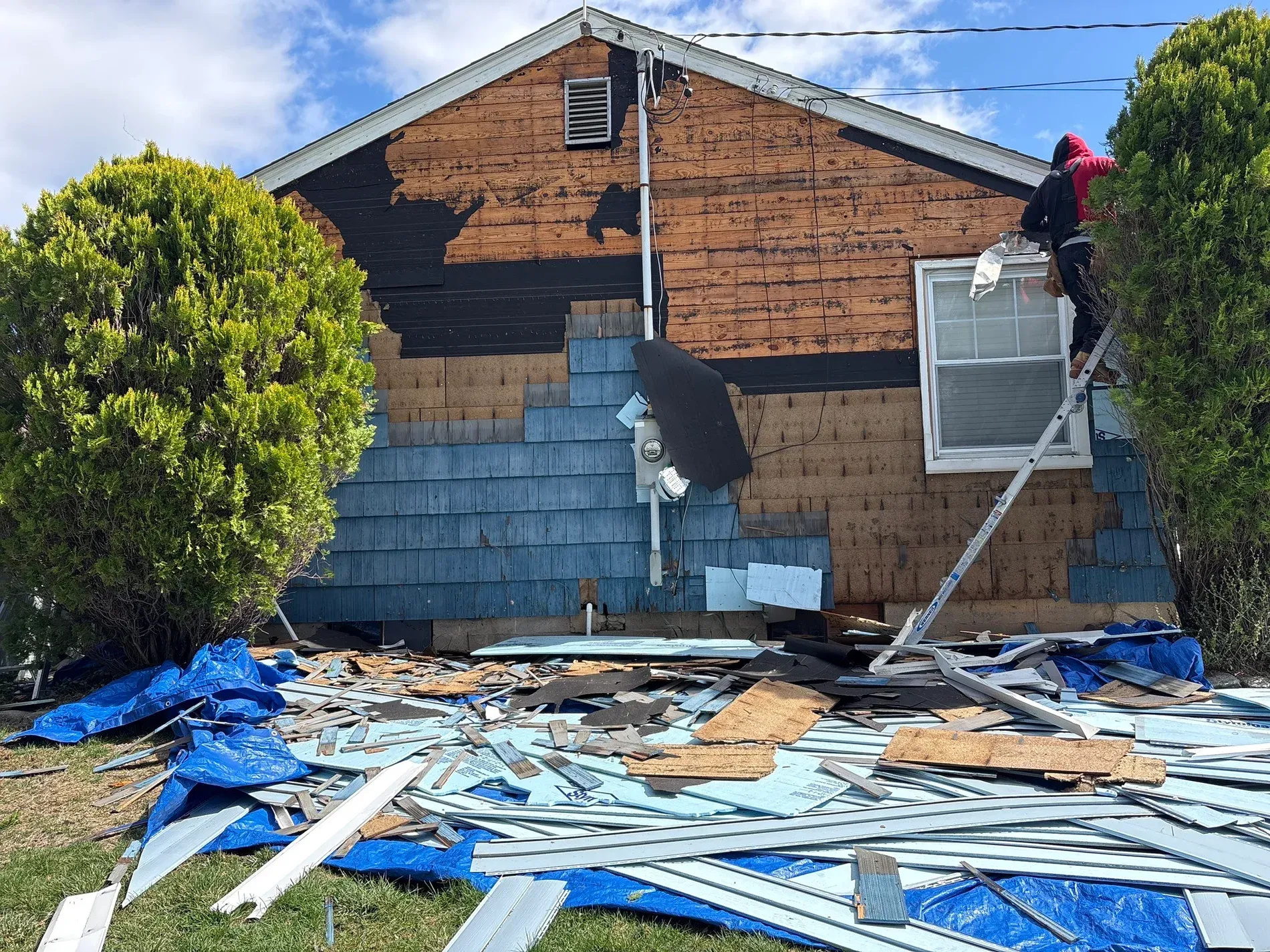Two workers repairing damaged house siding and roof, with debris scattered in the yard