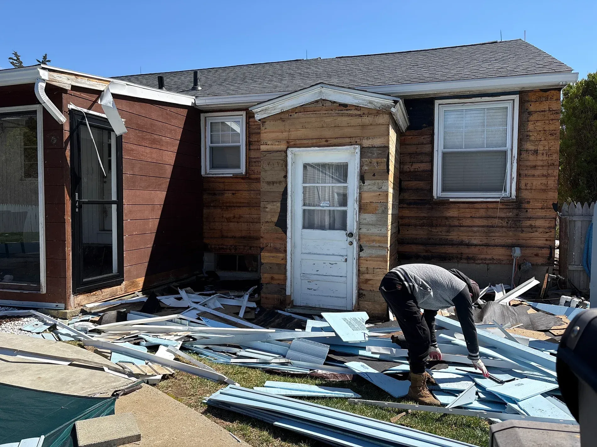 Worker amid debris outside a damaged brick house with a collapsed wall and broken roof