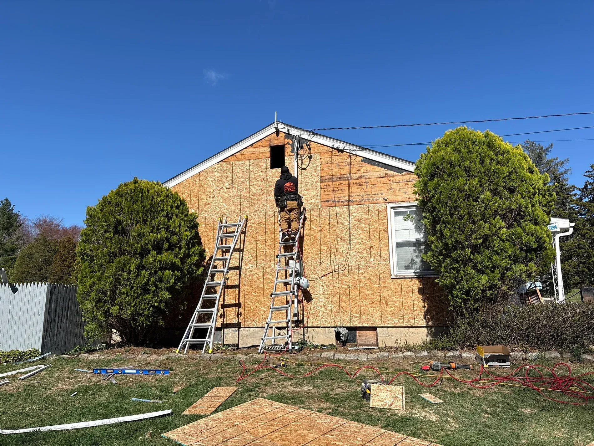 Brick house under repair with ladders and a worker on the roof, surrounded by trees and a grassy yard