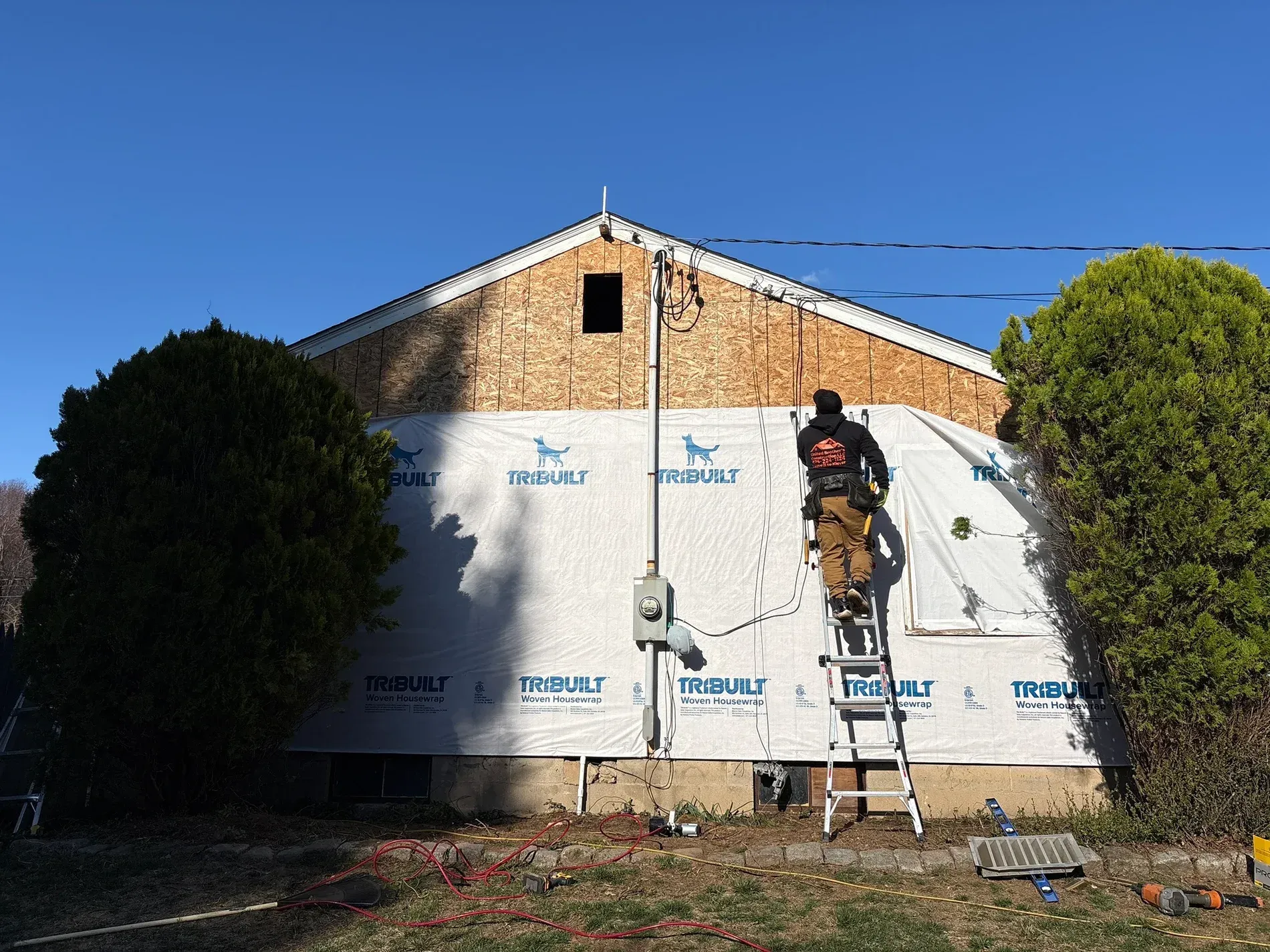 Worker on ladder installing white house wrap on a brick building under a clear blue sky
