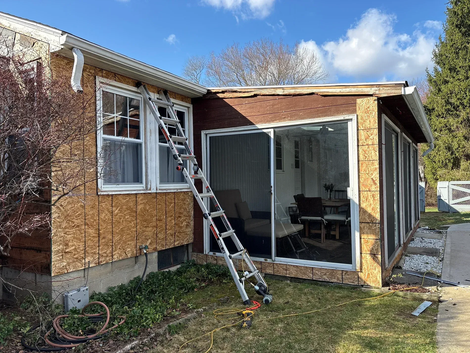 Exterior of a house with a ladder leaning against boarded-up windows and a screened porch under repair