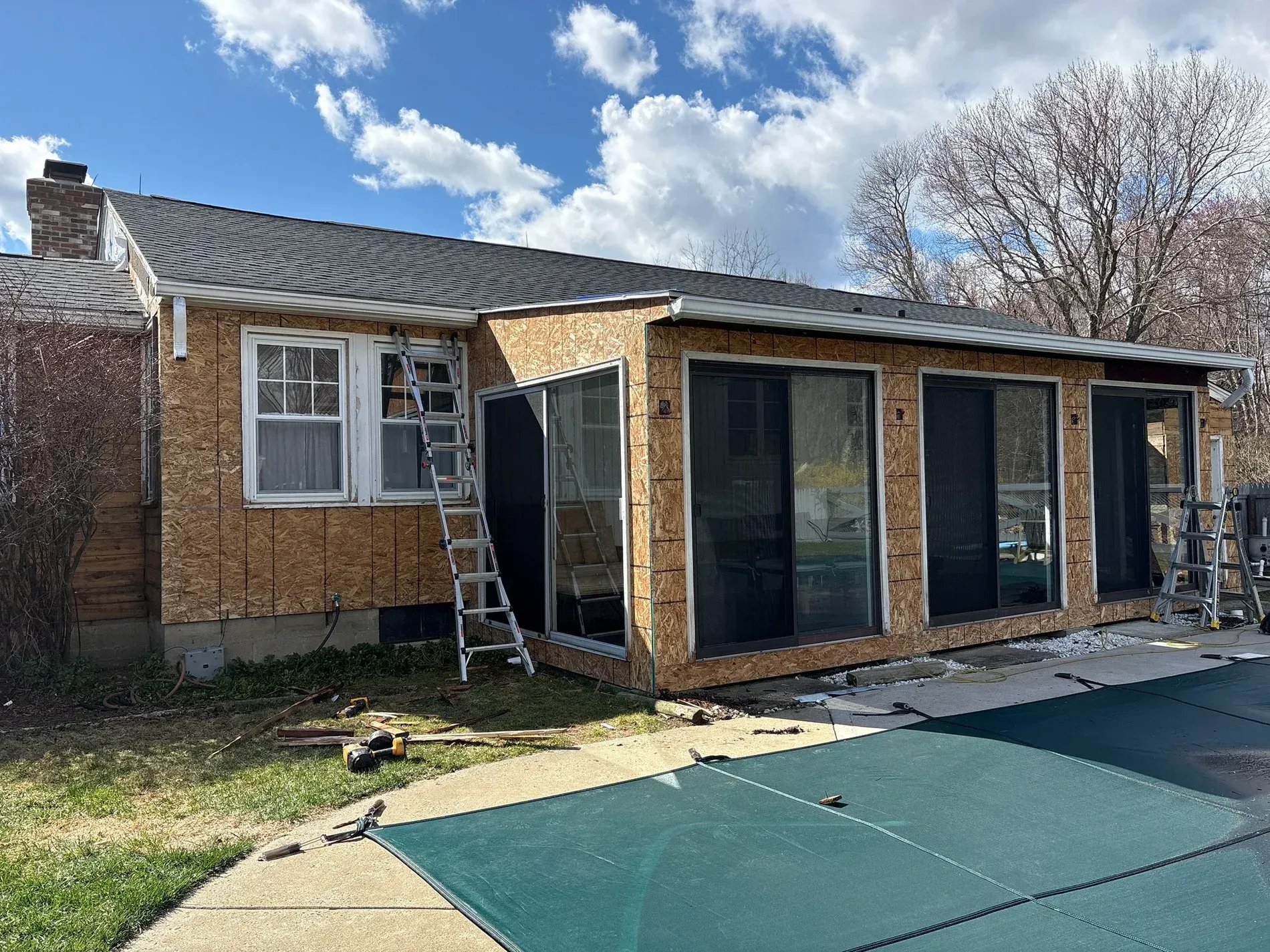 Backyard patio enclosure under construction beside a brick house, with black-framed glass doors and green flooring