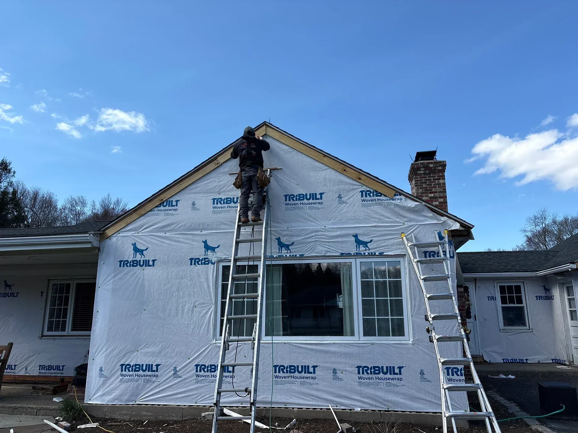 House under renovation with white house wrap, ladder, and worker on the roof under a blue sky