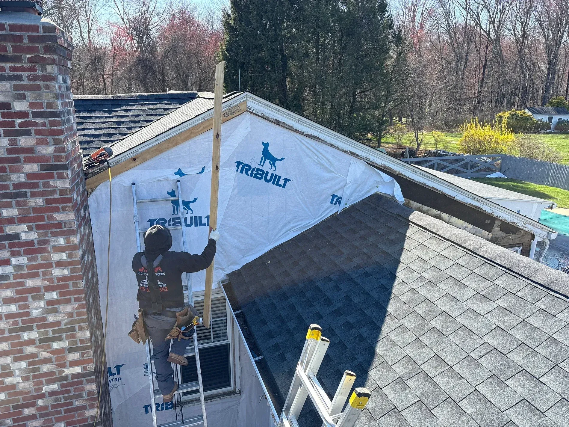 Worker installing roof underlayment on a house, with ladders and exposed roof framing visible.