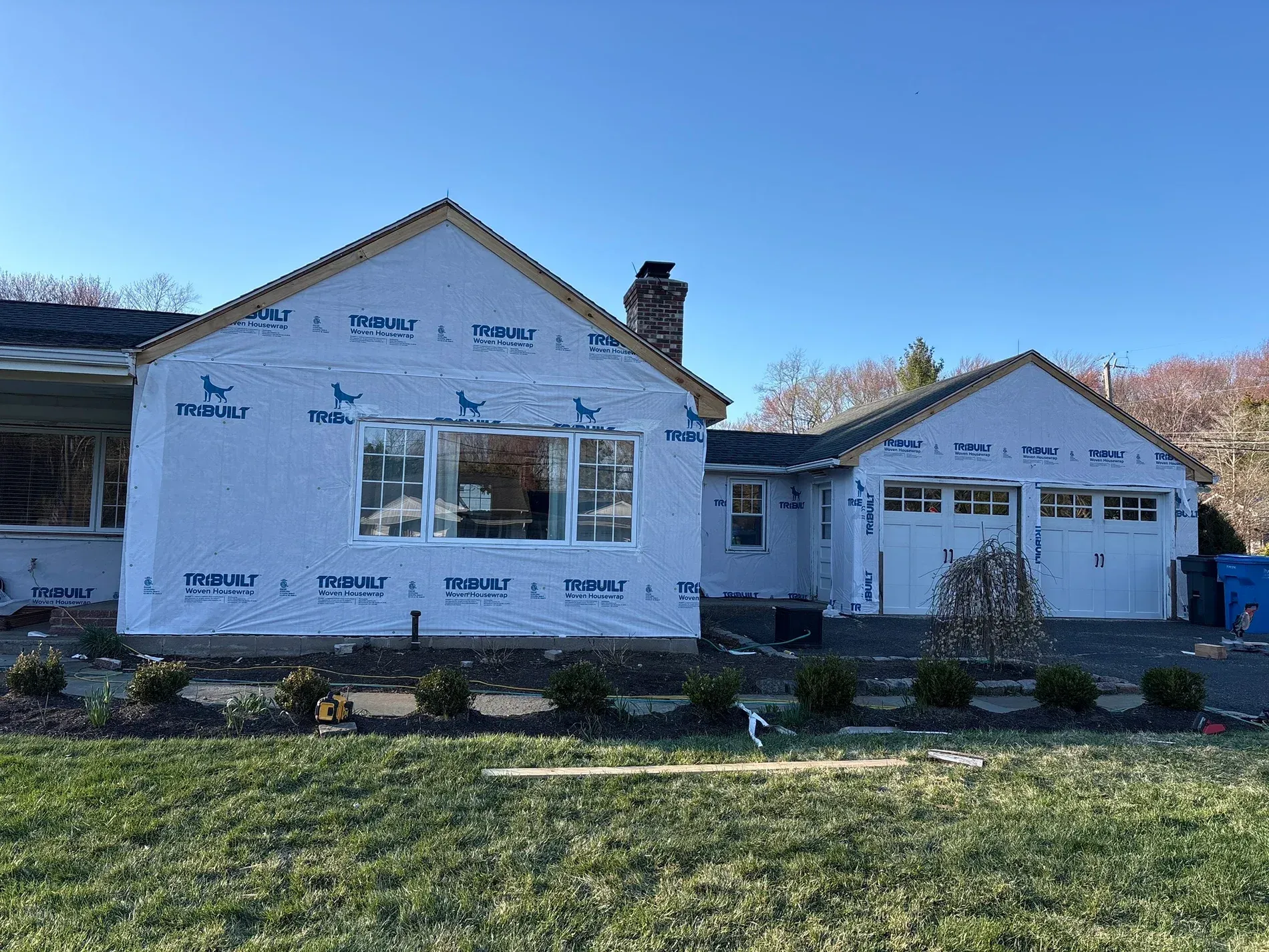 Partially remodeled suburban house with white siding wrap, garage, and landscaped front yard under blue sky